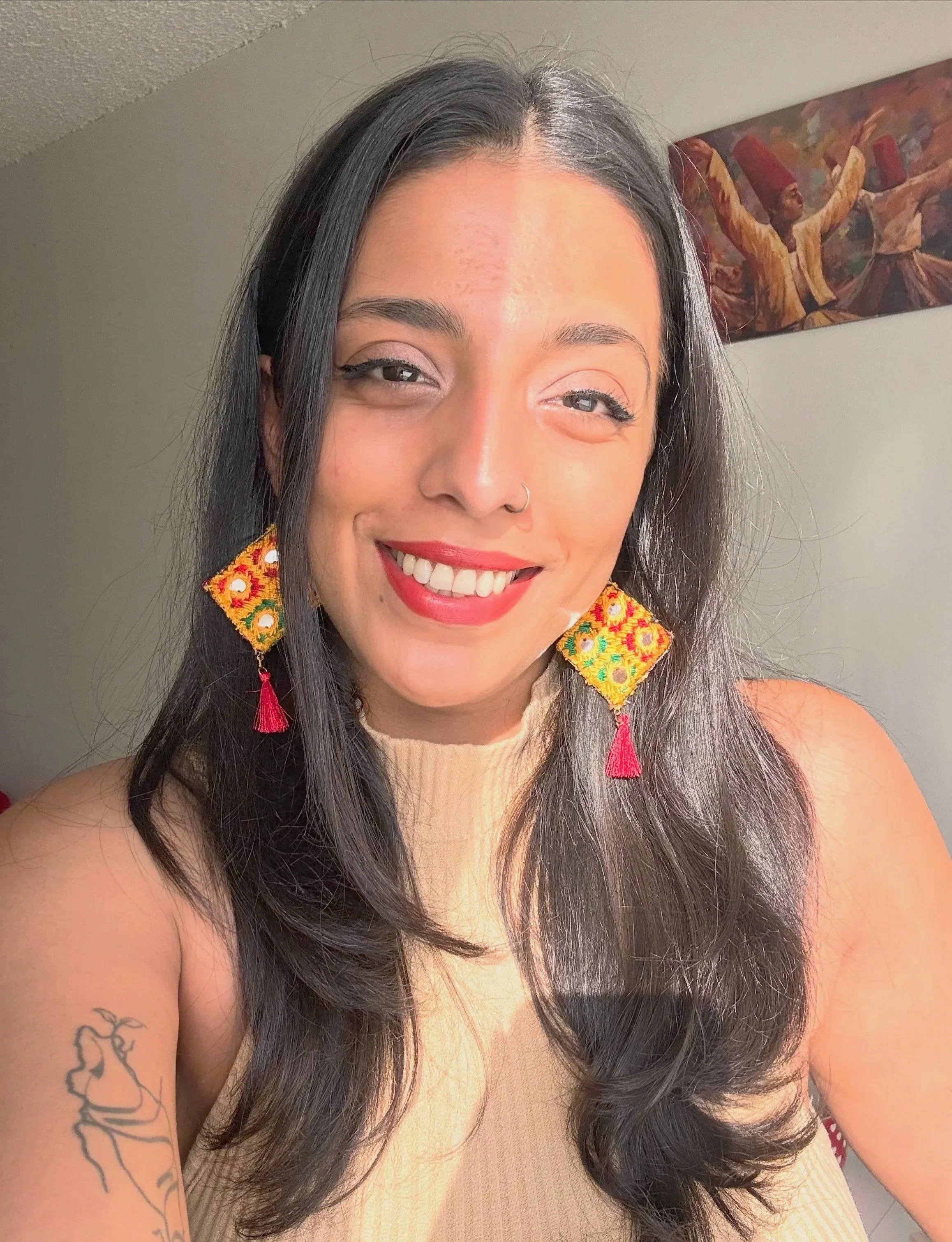 A young woman with long dark hair, light skin, and a nose ring smiling at the camera. She is wearing colorful embroidered earrings with pink tassels and a sleeveless beige top. There is a tattoo of a hand holding a rose on her upper arm. A painting with dancing figures is hanging on the wall behind her.