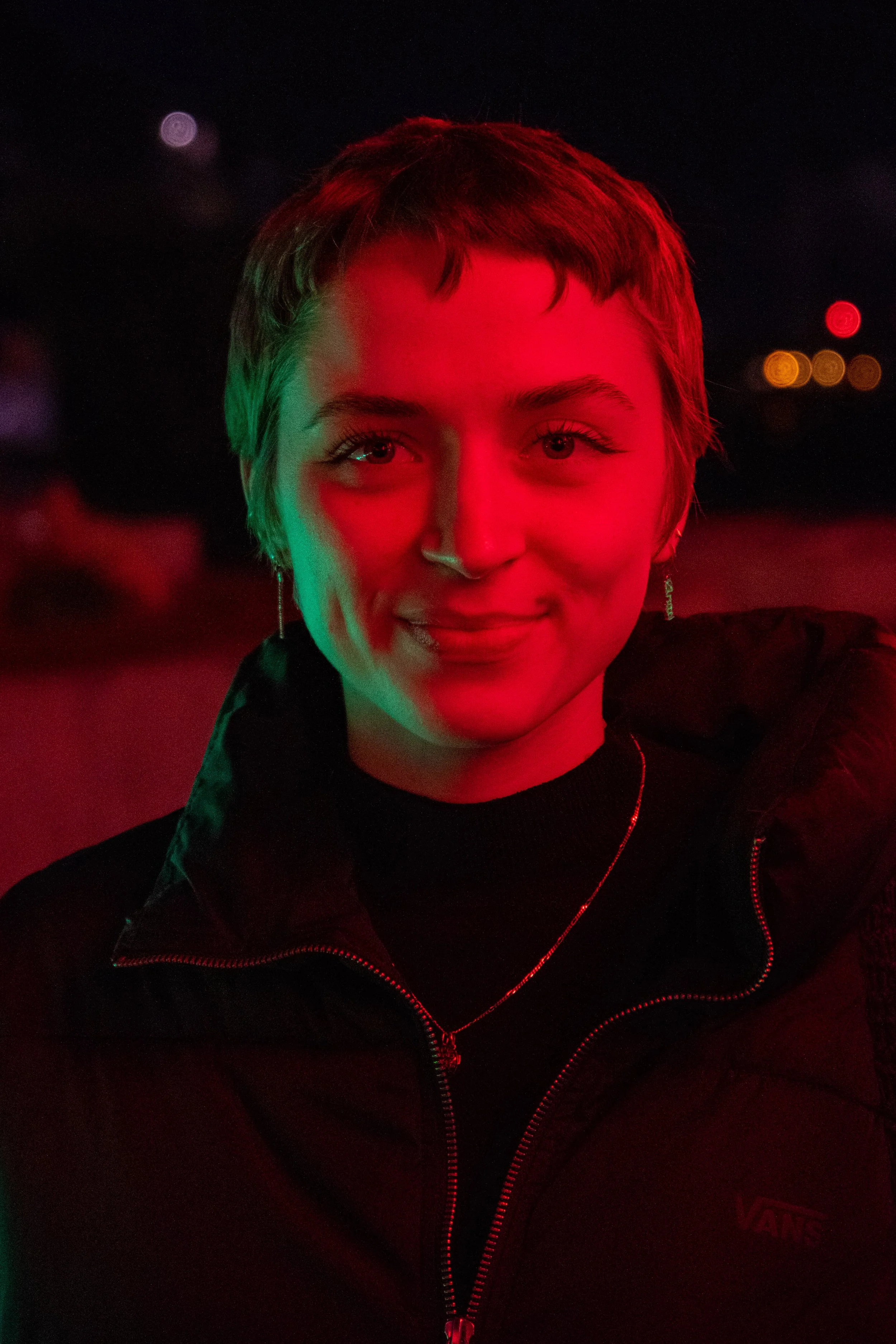 A person with short hair and fair skin illuminated with red and green lights at night, wearing a black jacket with a red zipper and earrings.