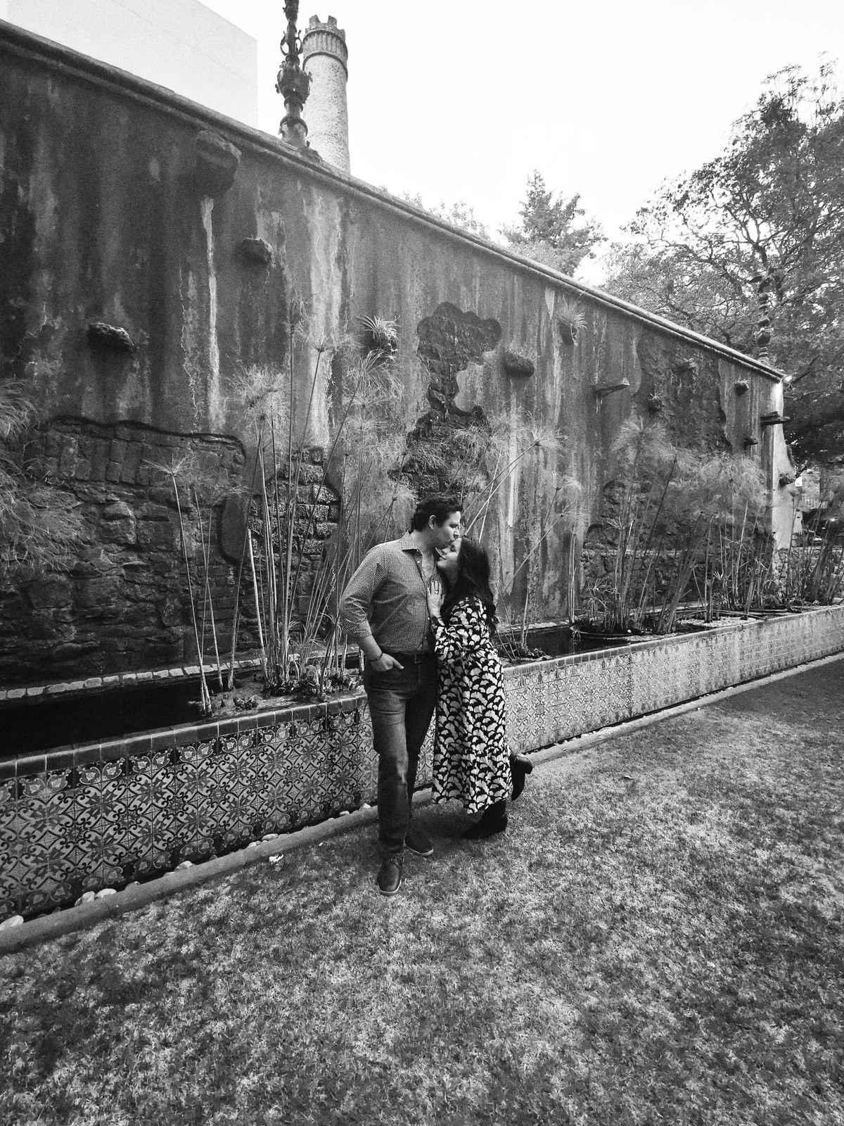 A black and white photo of a couple standing close together in front of a textured stone wall with plants growing along it. The man is facing right and the woman is leaning upward towards him, about to kiss. The scene is outdoors, possibly during the
