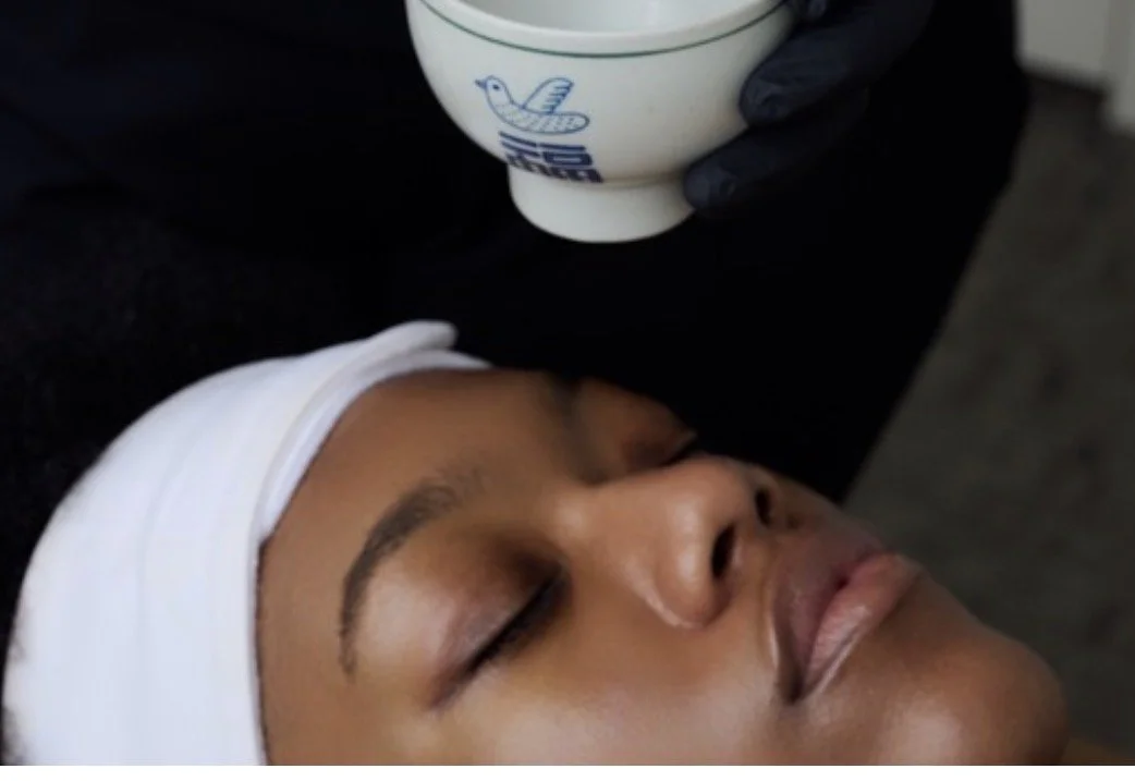 A woman receiving a facial massage, with her eyes closed, lying down with a white towel wrapped around her head.
