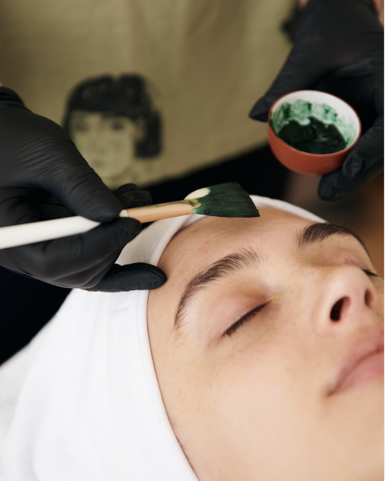 A woman with black hair and earrings getting a hair treatment with a syringe from a professional wearing a beige apron and black gloves. The background shows a sign saying 'SORA skin + scar' on a white wall.