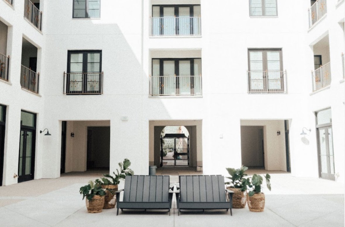 Courtyard of a modern apartment building with white walls, black-framed windows, and balconies. There are two black outdoor chairs and potted plants in woven baskets in the center.