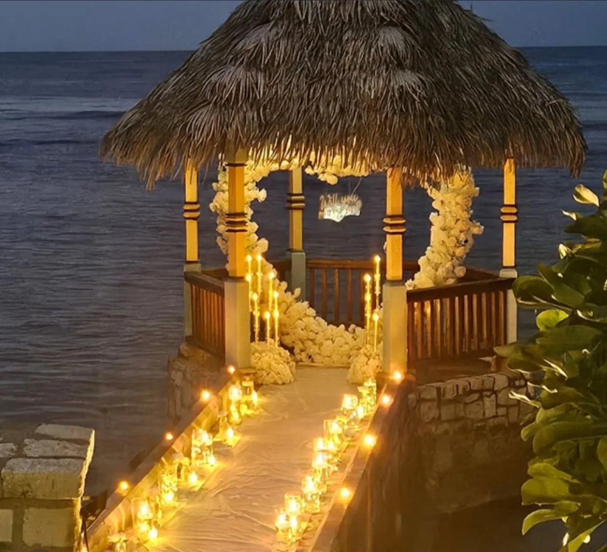 A romantic wedding altar on a pier over the water with a thatched roof, floral decorations, and candles lining the aisle.