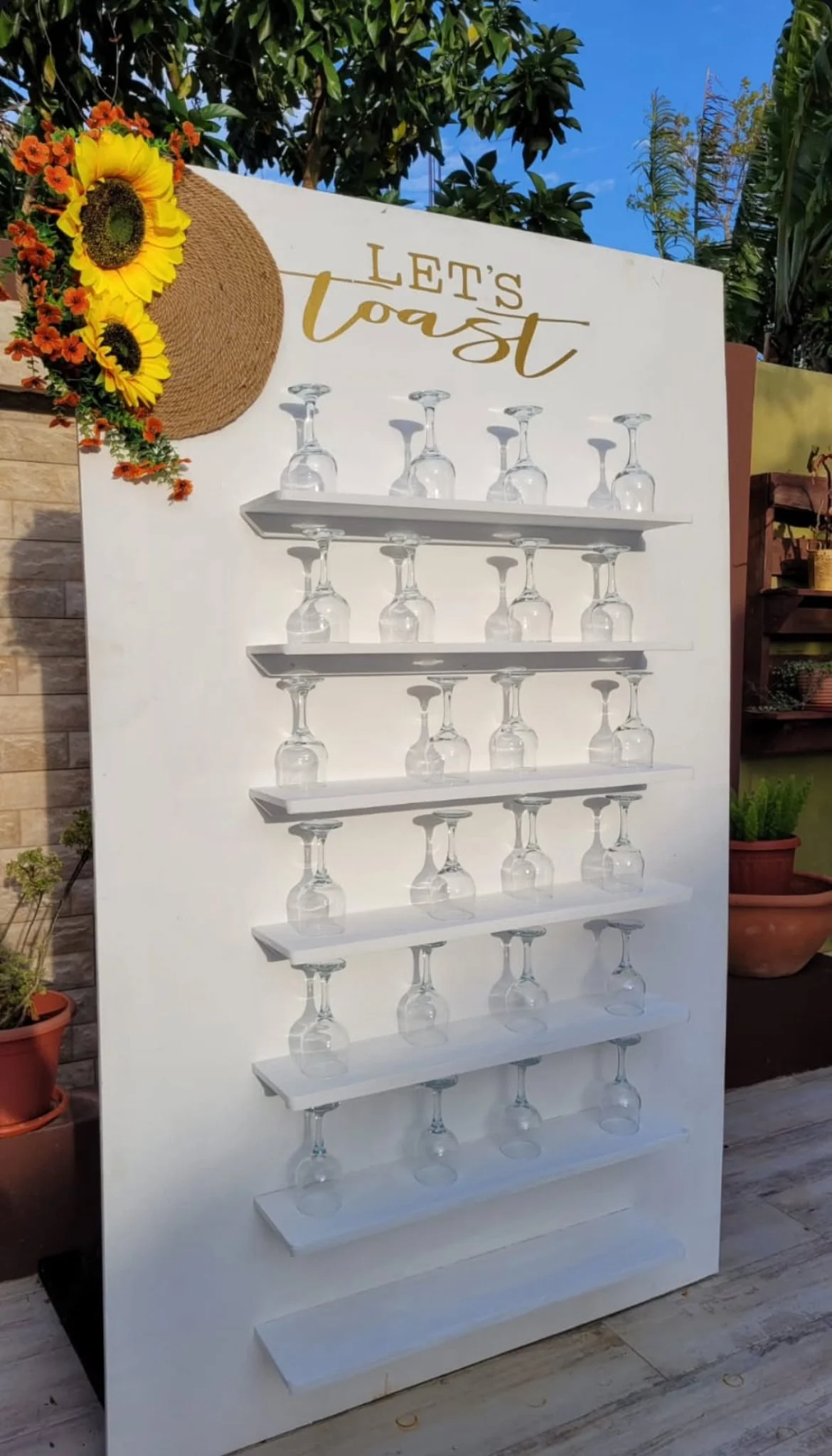 A white display stand with wine glasses upside down on it, situated outdoors with plants and a blue sky in the background. The top of the stand has a sentiment reading 'Let's Toast' in gold lettering, decorated with a sunflower and a straw hat.