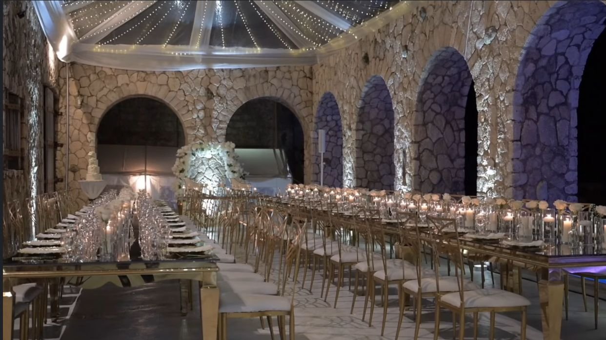 Elegant indoor event space with a long dining table, gold chairs, and candle lighting, featuring a stone wall and arched windows in the background.