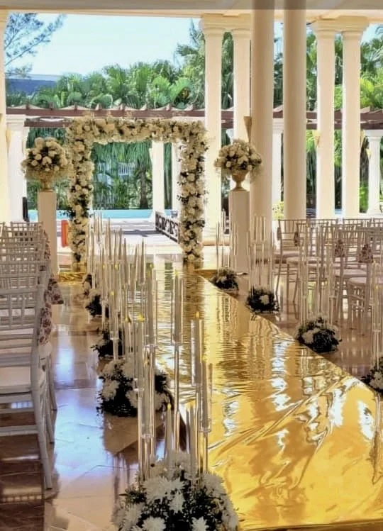 Wedding aisle with white flowers and chairs on sides under a decorated canopy with greenery in an outdoor setting.