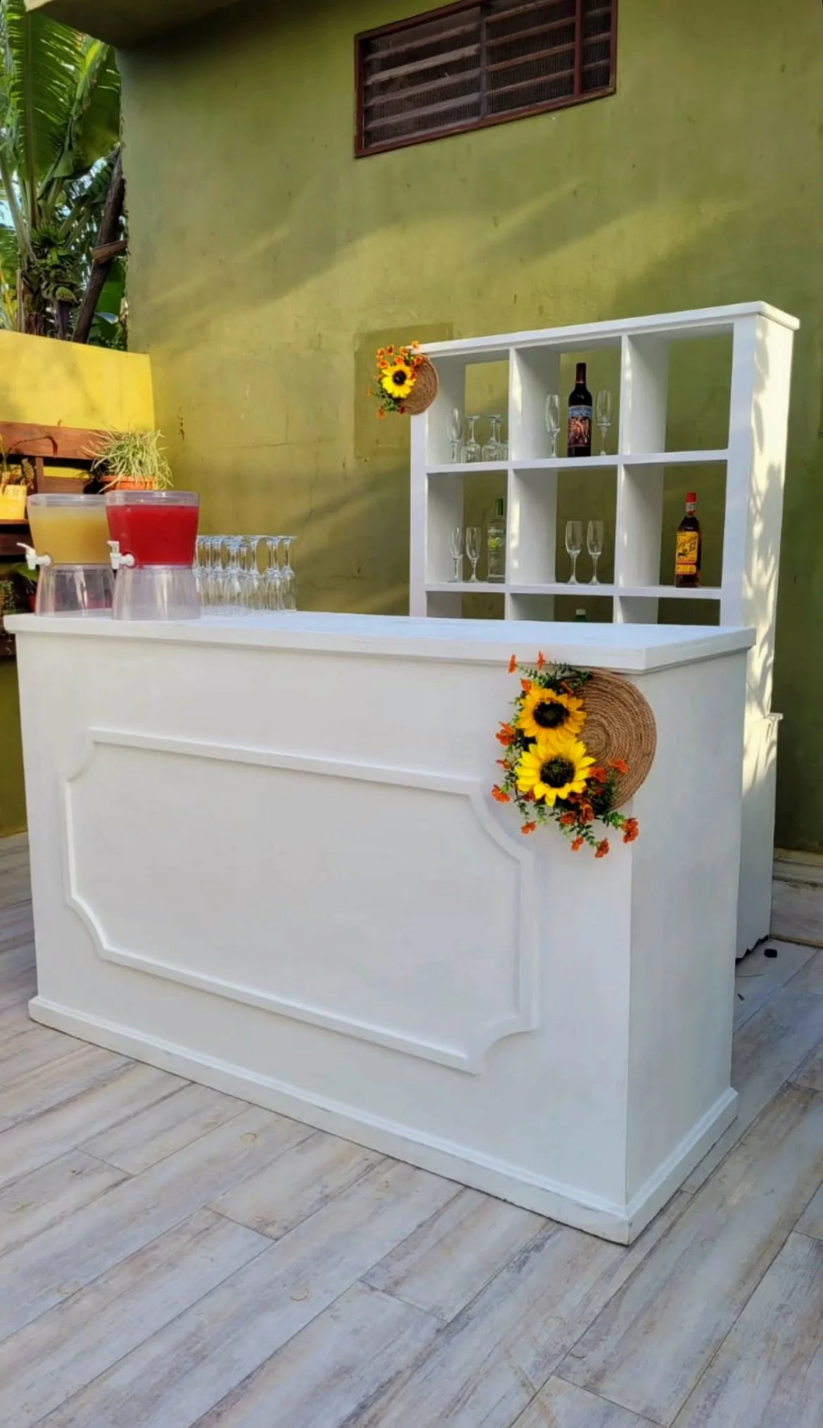 A white bar counter with sunflower decorations on the side, set outdoors against a green wall with a window. The counter has bottles, glasses, and drink dispensers on top, and a white shelving unit behind it holds liquor bottles and glassware.