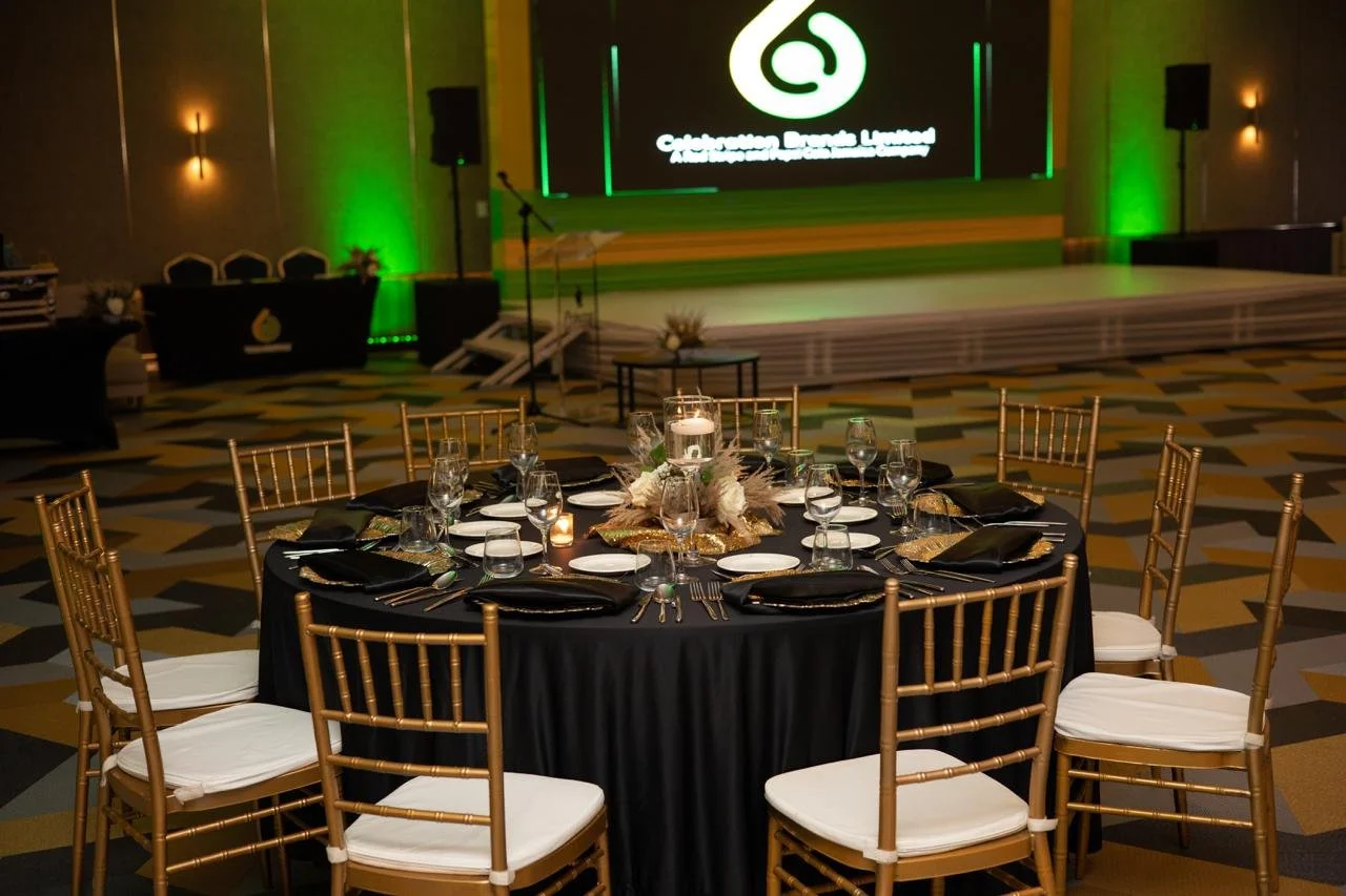 Round banquet table with black tablecloth, gold chairs with white cushions, and a centerpiece with candles and feathers, set for a formal event in a decorated ballroom with a stage and digital screen in the background.