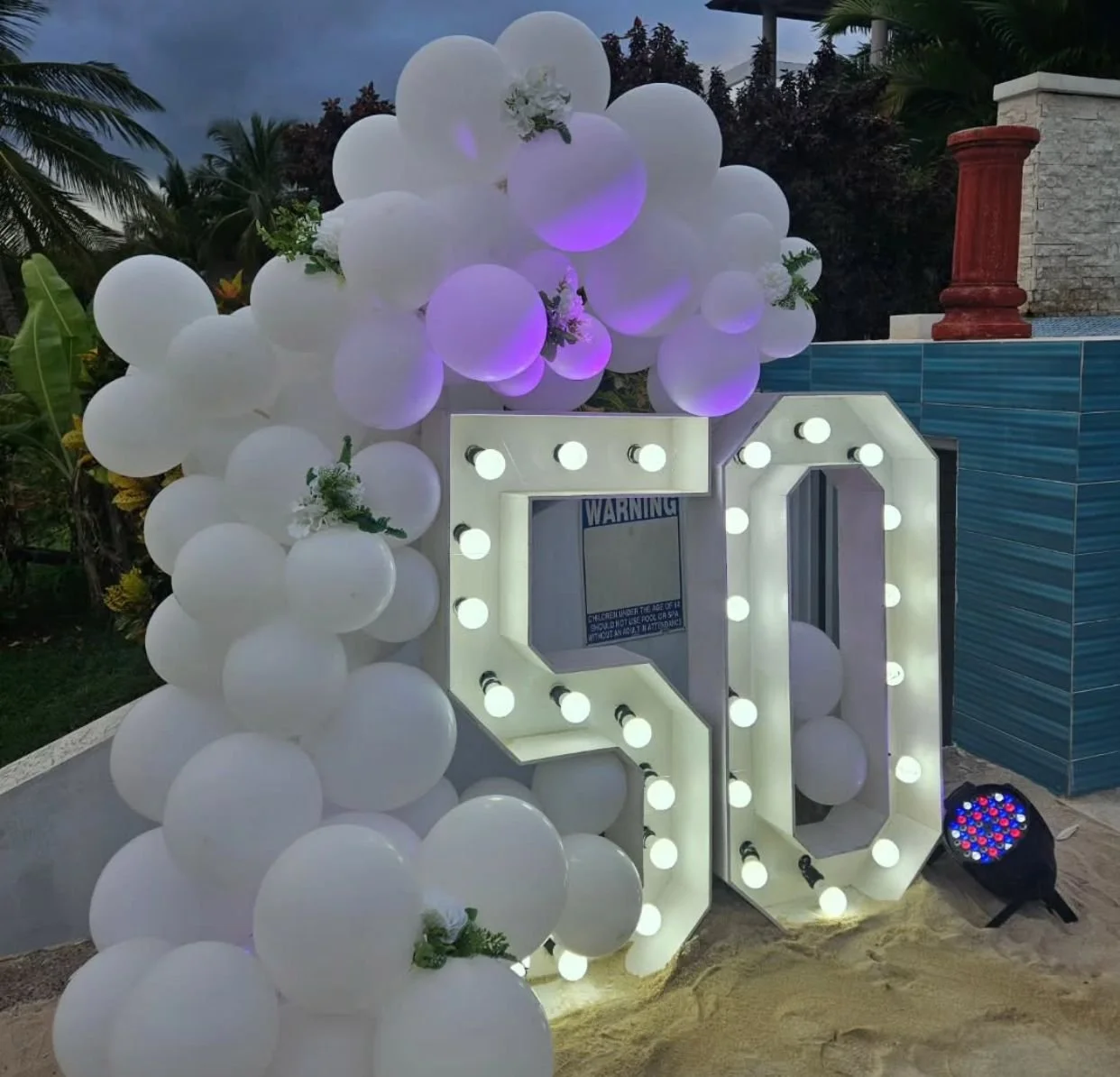 Decorative setup with white balloons, illuminated marquee letters, and purple lighting, outdoors at dusk with tropical plants and a pool area in the background.