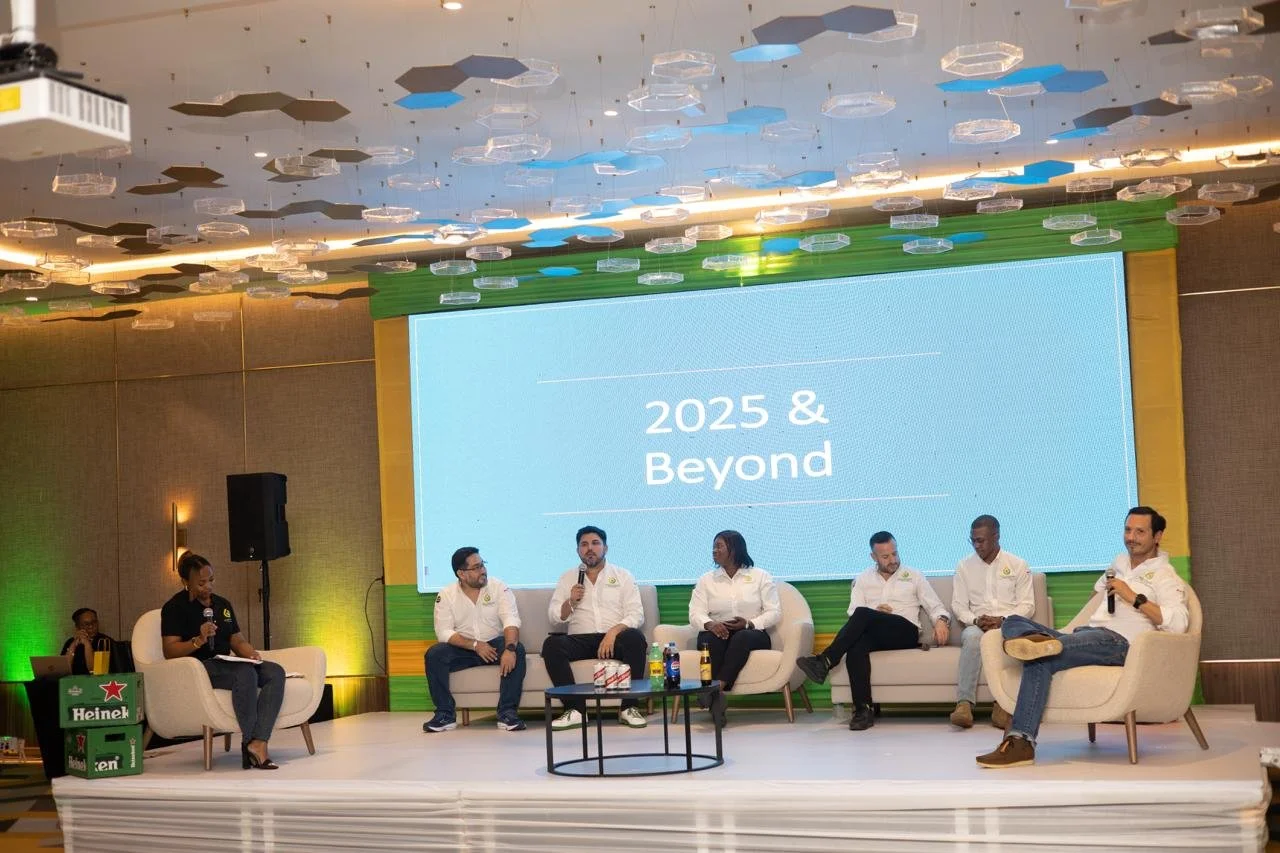 A panel discussion with seven individuals on stage, five seated on a sofa and two on individual chairs, with a large screen behind displaying '2025 & Beyond', in a conference room with hexagonal ceiling lights.