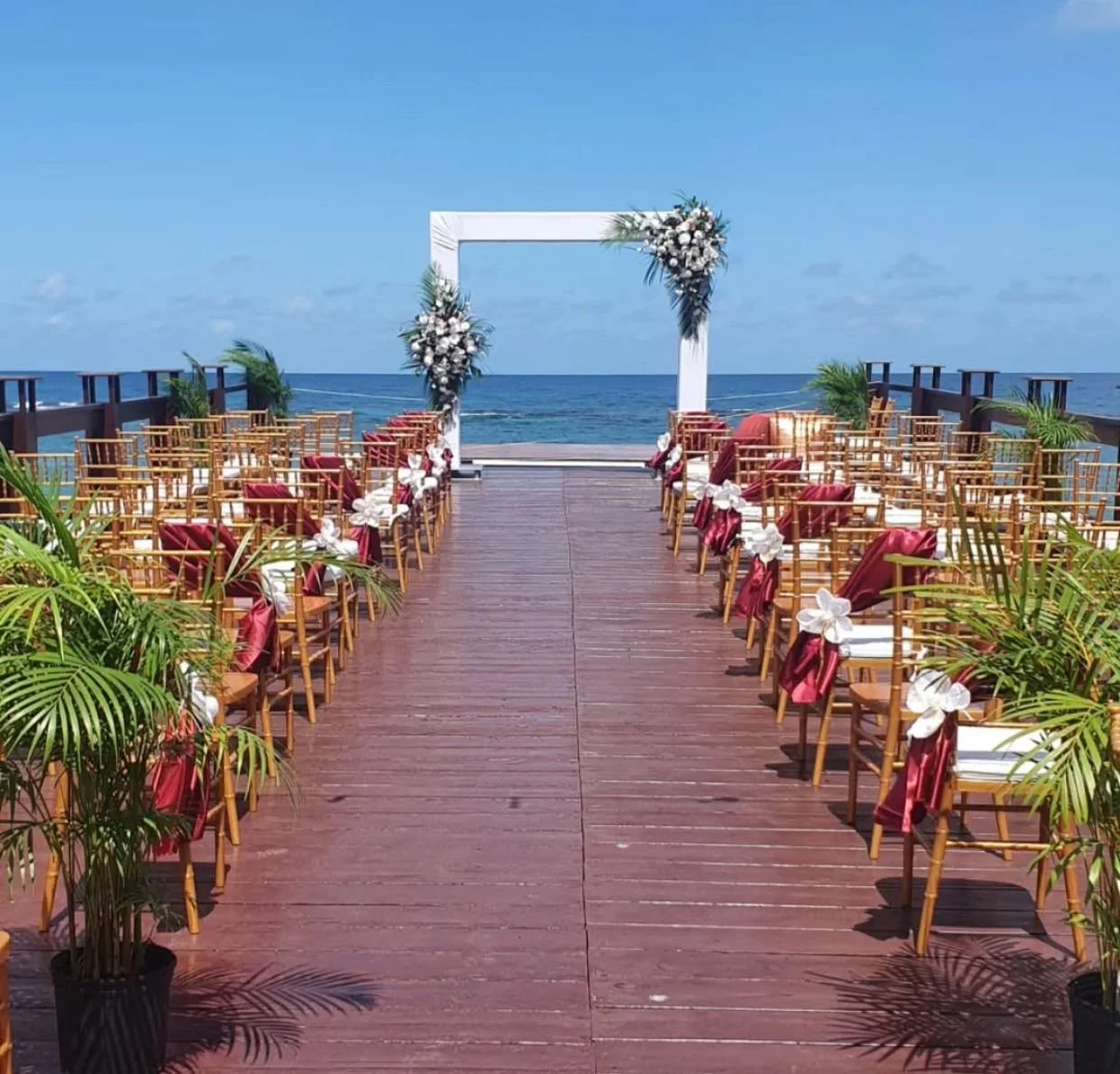 Wedding arch setup on a wooden pier by the ocean with chairs decorated with burgundy ribbons and potted green plants.