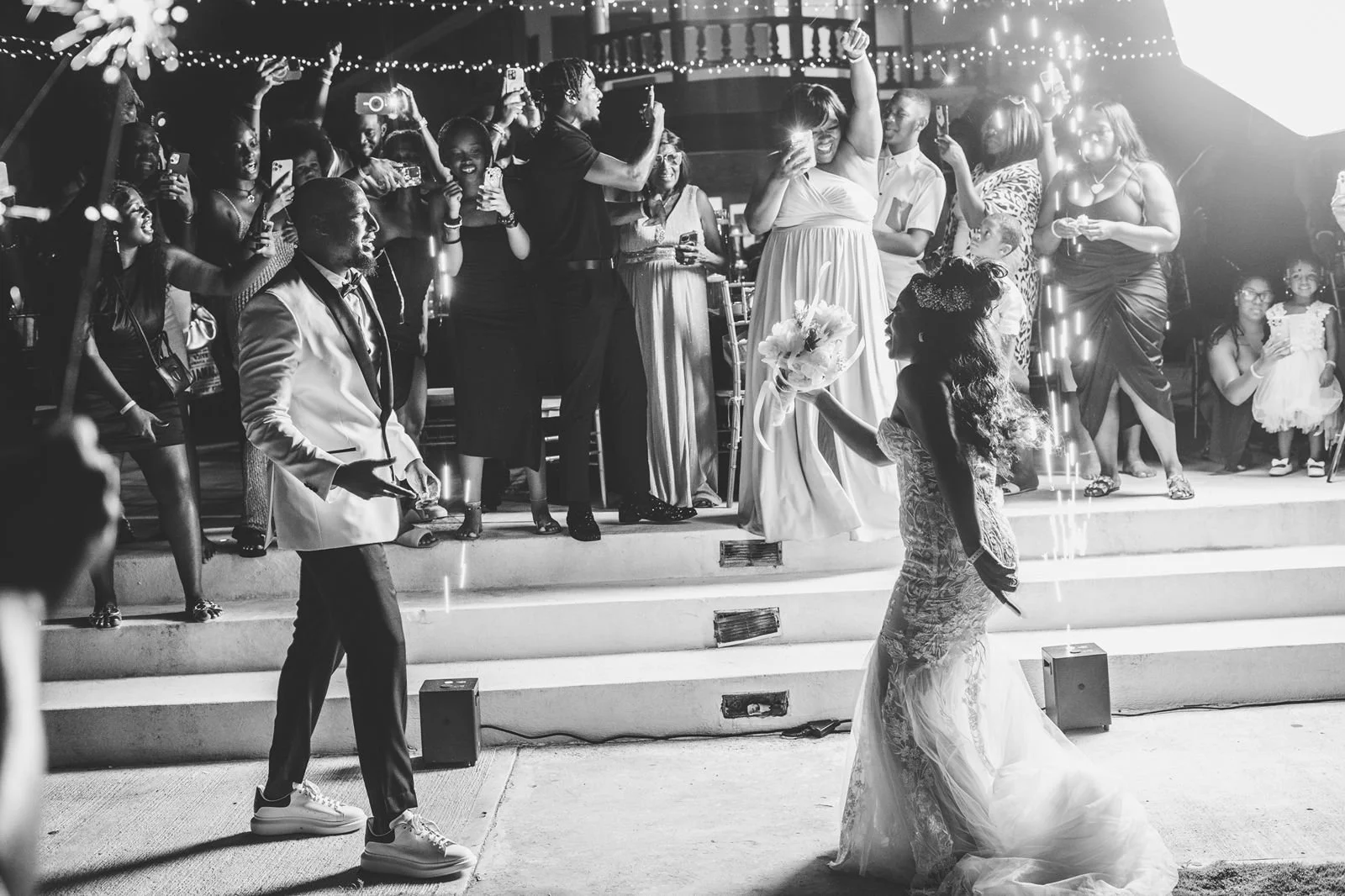 Wedding reception with bride, groom, and guests, some taking photos or videos, in a decorated venue with string lights.