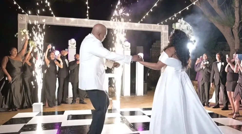 A bride and groom dancing at their wedding reception, with guests cheering and celebrating in the background, night sky, and sparklers.