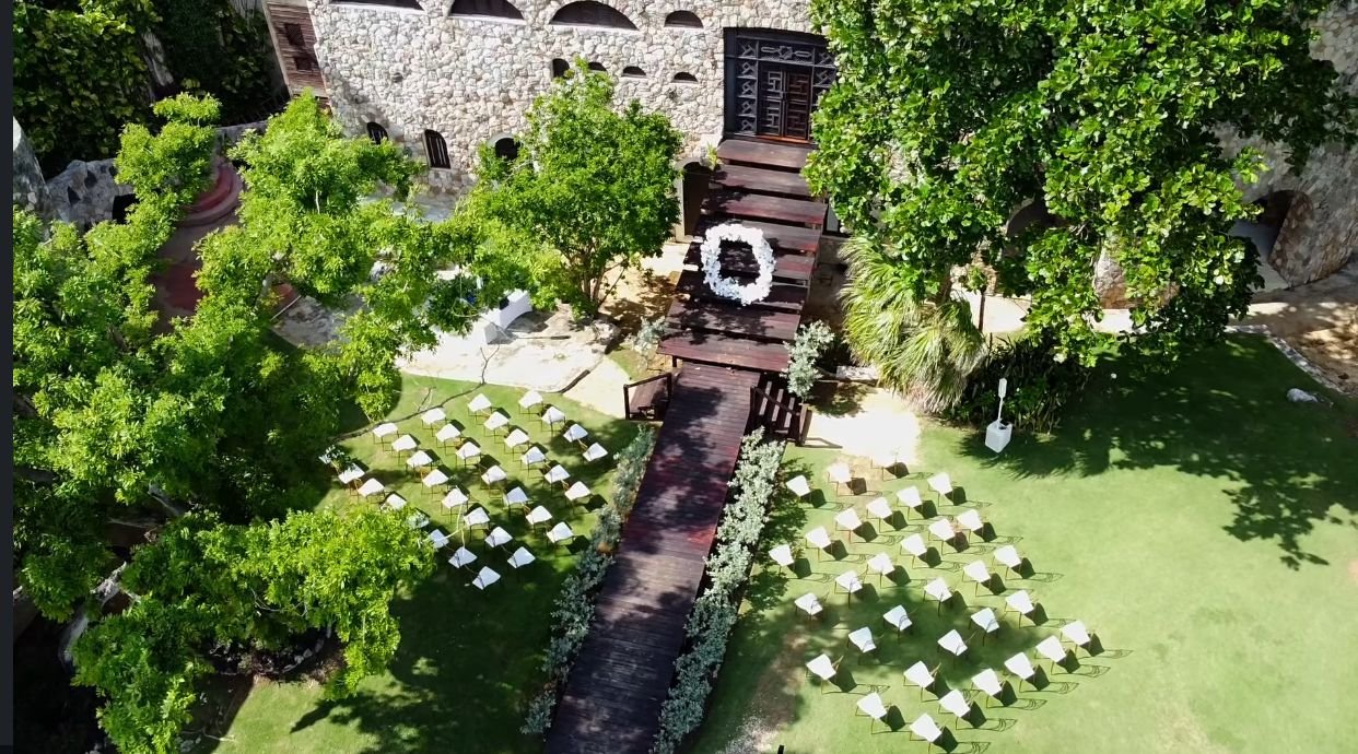 Aerial view of an outdoor wedding setup with white chairs arranged in rows on grass, a wooden pathway leading to a building with a wreath, and trees providing shade.