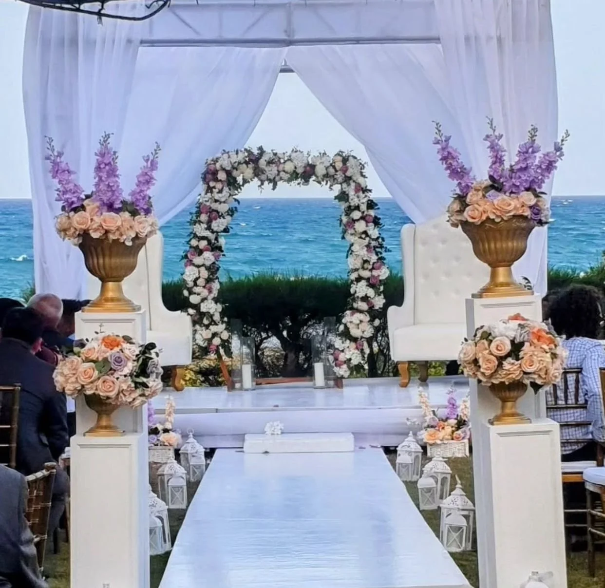 Beachfront wedding arch decorated with white curtains and floral arrangements, with chairs on either side, set up for a ceremony overlooking the ocean.
