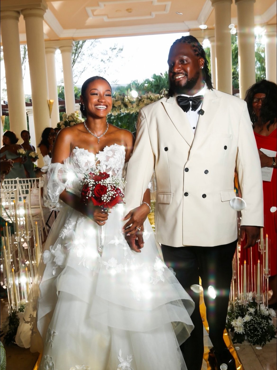 A smiling bride and groom holding hands during their wedding ceremony, with the bride holding a bouquet of red roses, inside a decorated venue with guests in the background.