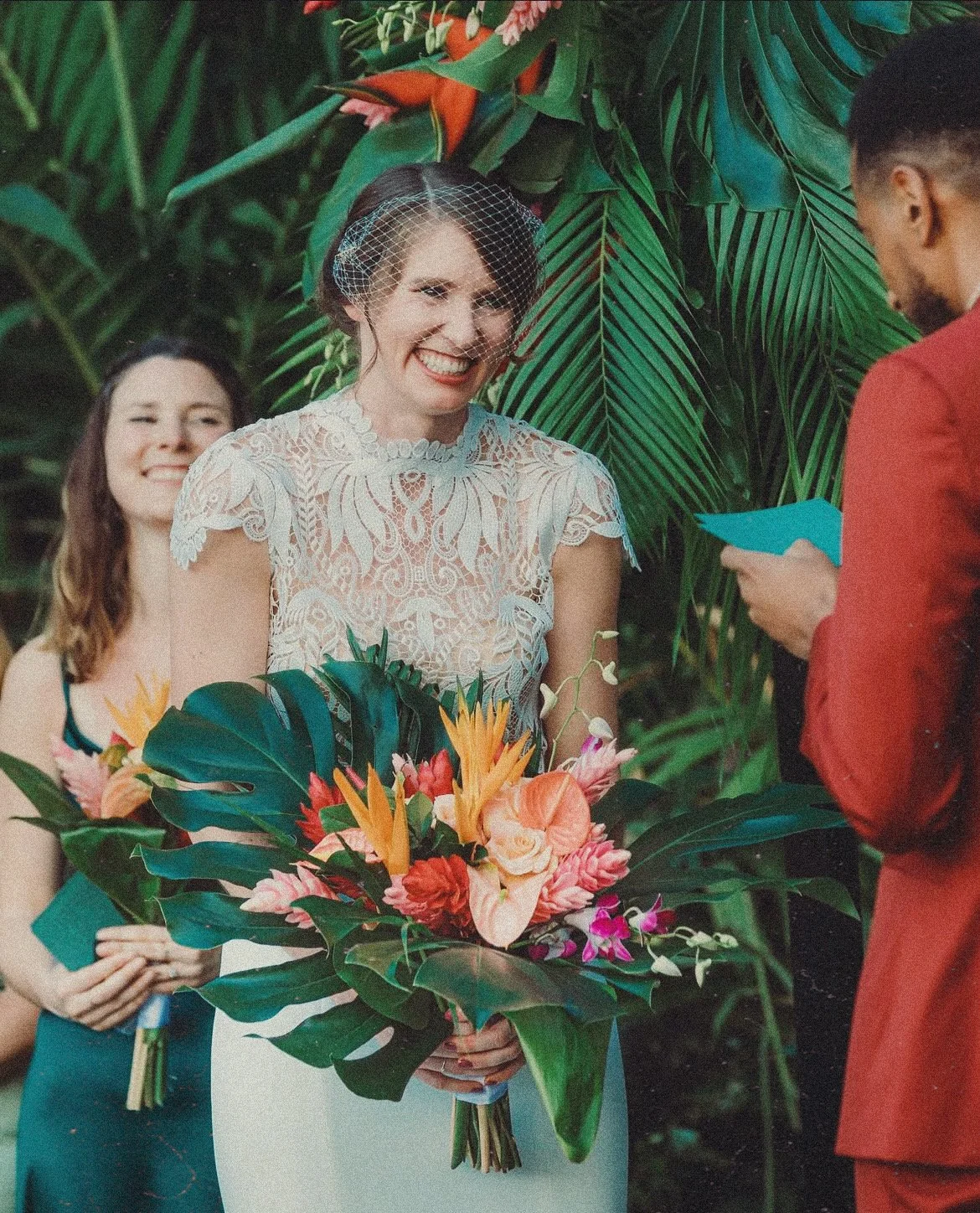 A woman in a white lace dress holding a tropical floral bouquet during a wedding ceremony, smiling at an officiant, with a bridesmaid holding a smaller bouquet in the background, all surrounded by lush green tropical plants.
