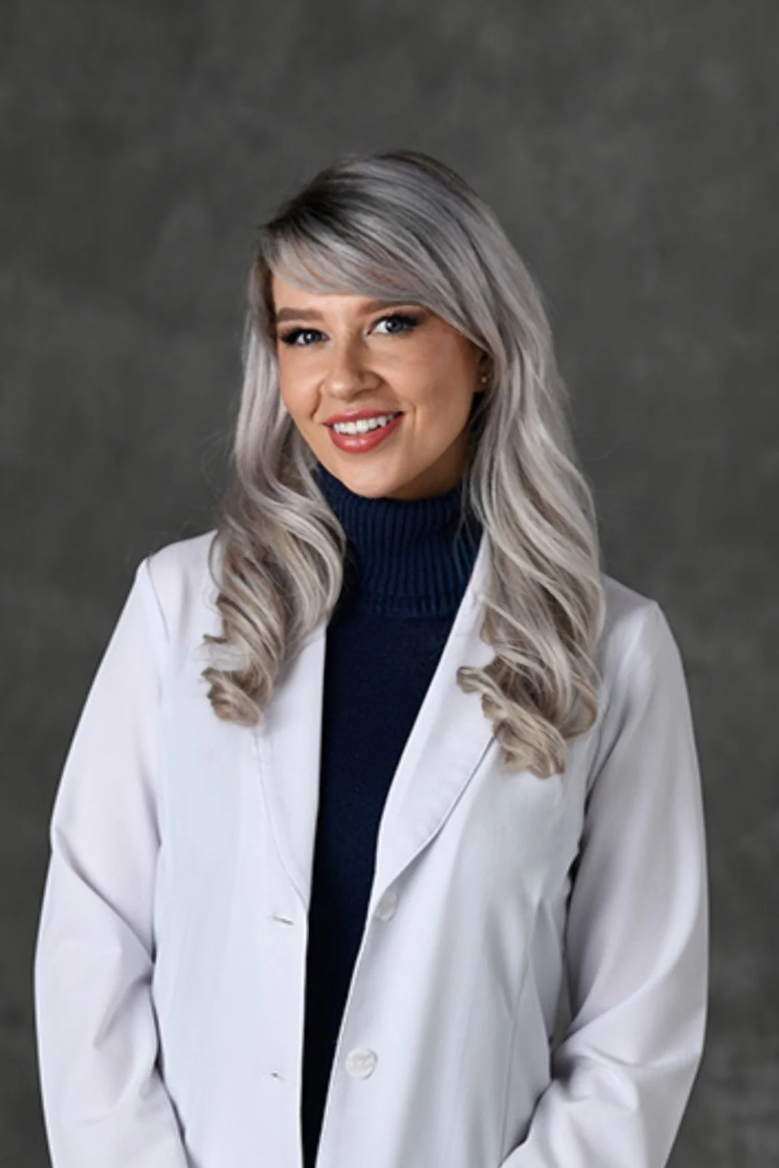 A smiling woman with long, wavy, silver-gray hair wearing a white coat over a dark blue turtleneck, standing against a gray background.