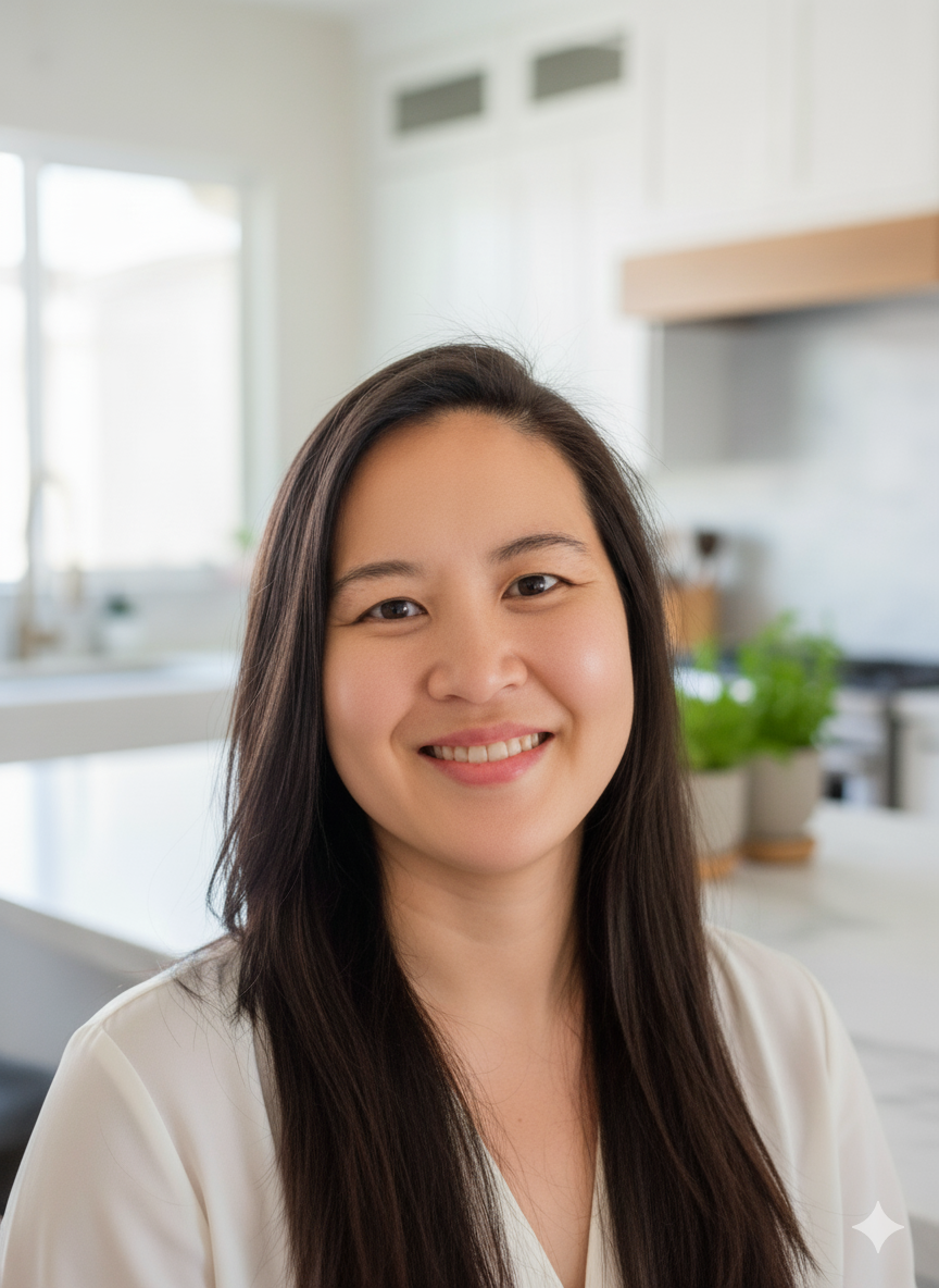 Portrait of a smiling Asian woman with long dark hair, standing in a bright, modern kitchen with white cabinets and windows in the background.