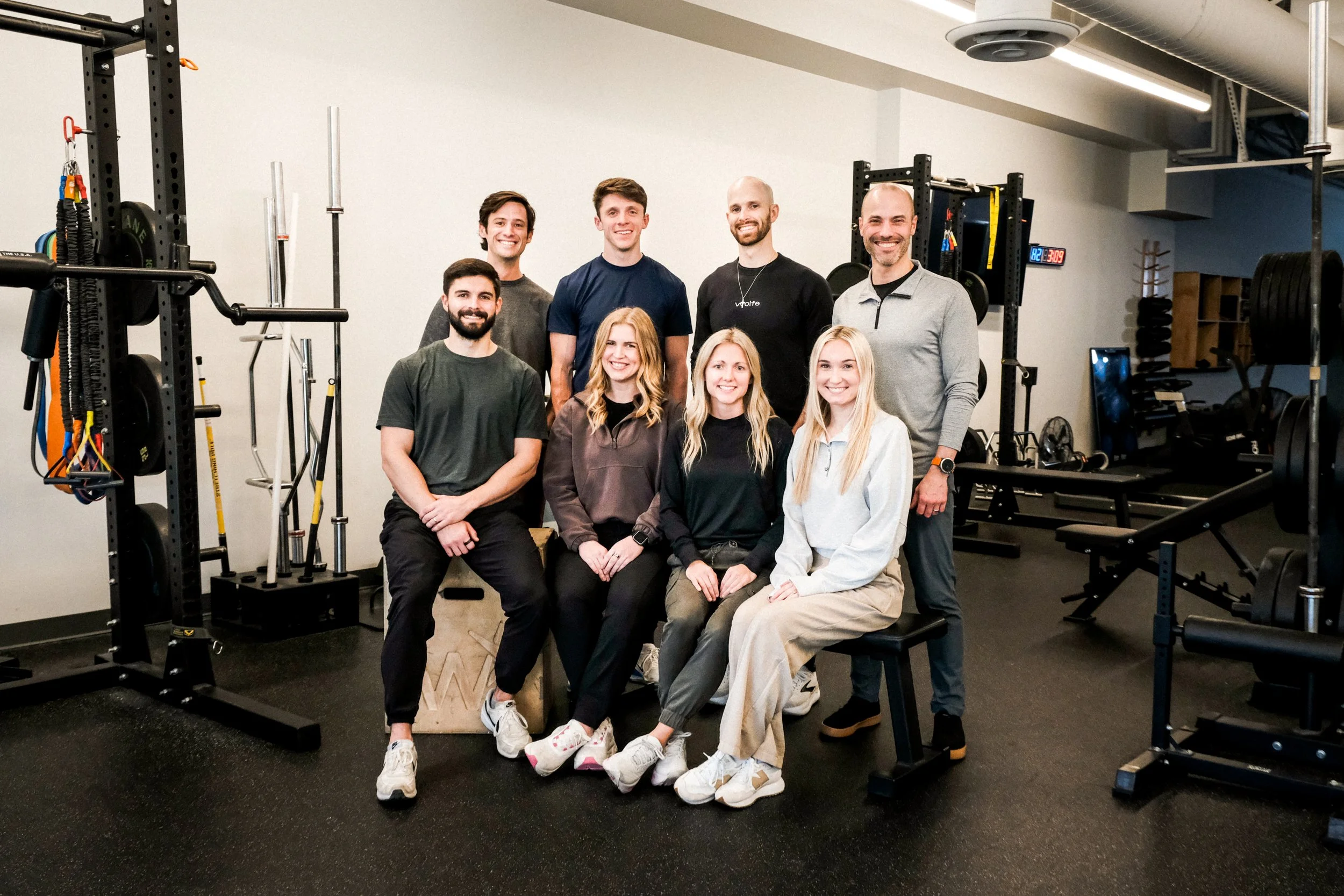 Group of nine people in a fitness gym posing for a photo, with gym equipment visible in the background.