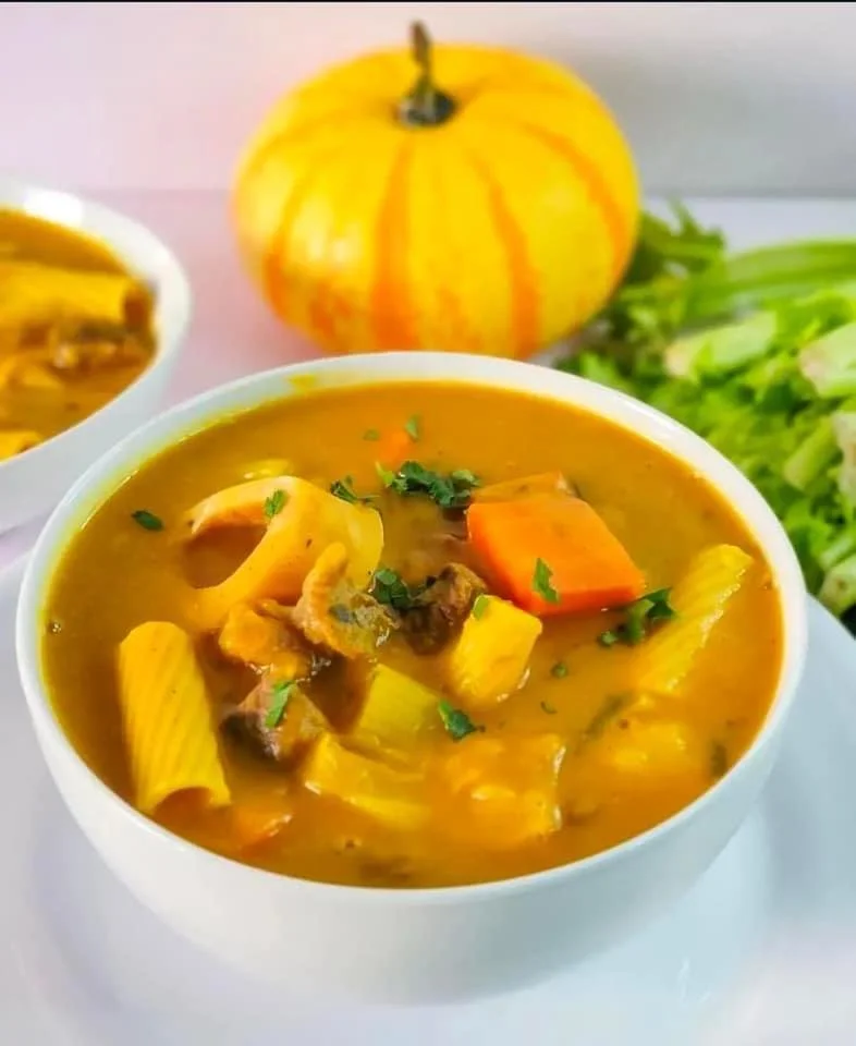 A bowl of vegetable soup with pasta, carrots, and herbs, with a small pumpkin in the background and fresh greens to the side.