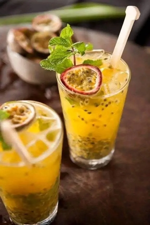 Two glasses of passion fruit lemonade garnished with passion fruit slices, lemon slices, mint leaves, and a straw, on a dark wooden table.