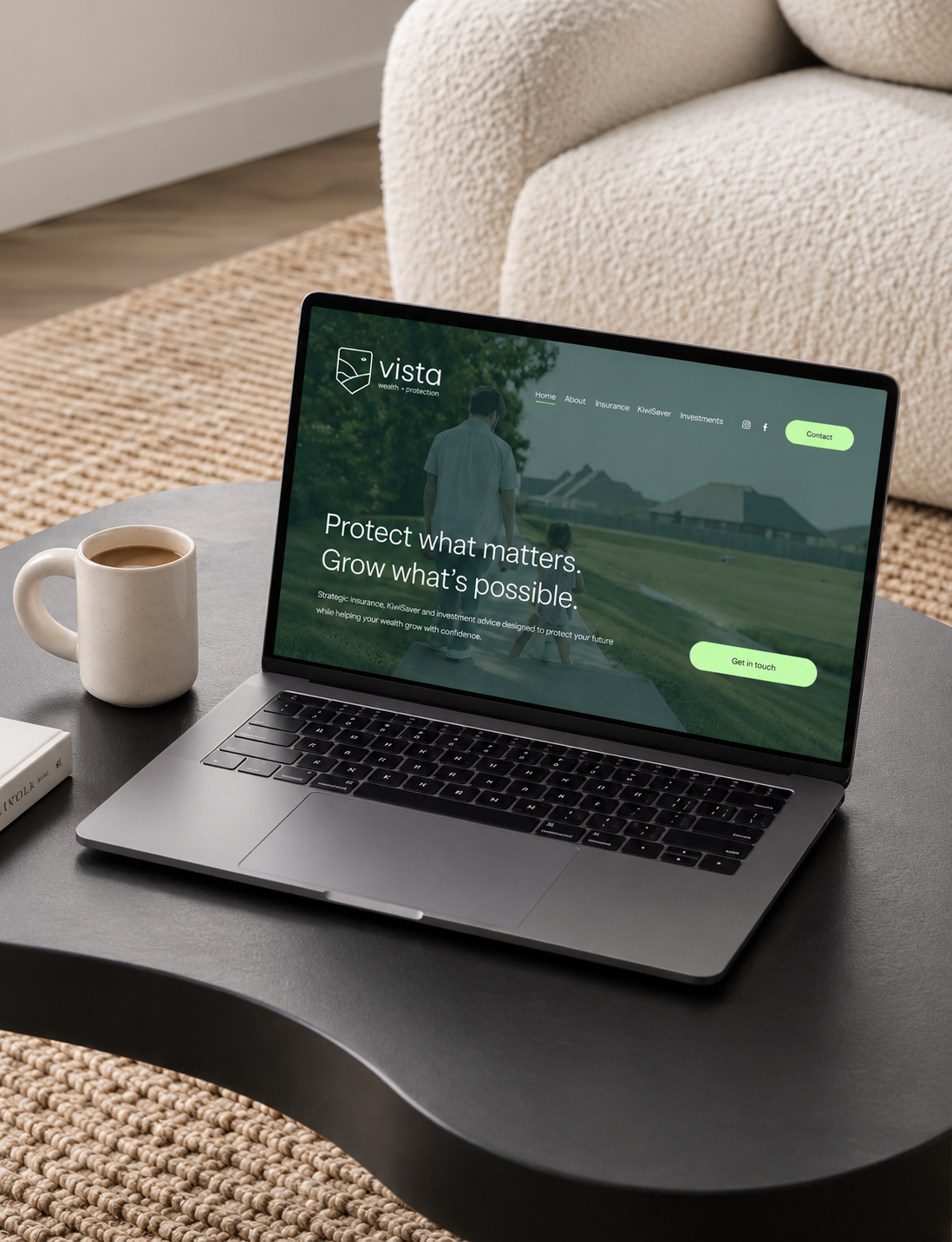 A laptop on a black table displays a website for Vista, an insurance and investment company, with the text 'Protect what matters. Grow what’s possible.' There is a cream-colored mug of coffee, a closed book, and a woven rug on the table. A beige armc