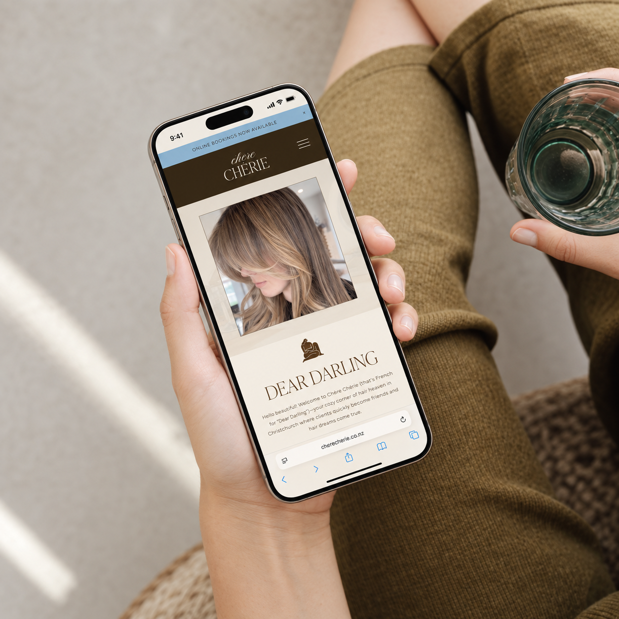 Person sitting on a woven chair holding a smartphone in their left hand, viewing a website for Chère Cherie hair salon with a photo of a woman with long, wavy hair, and a glass of water in their right hand.