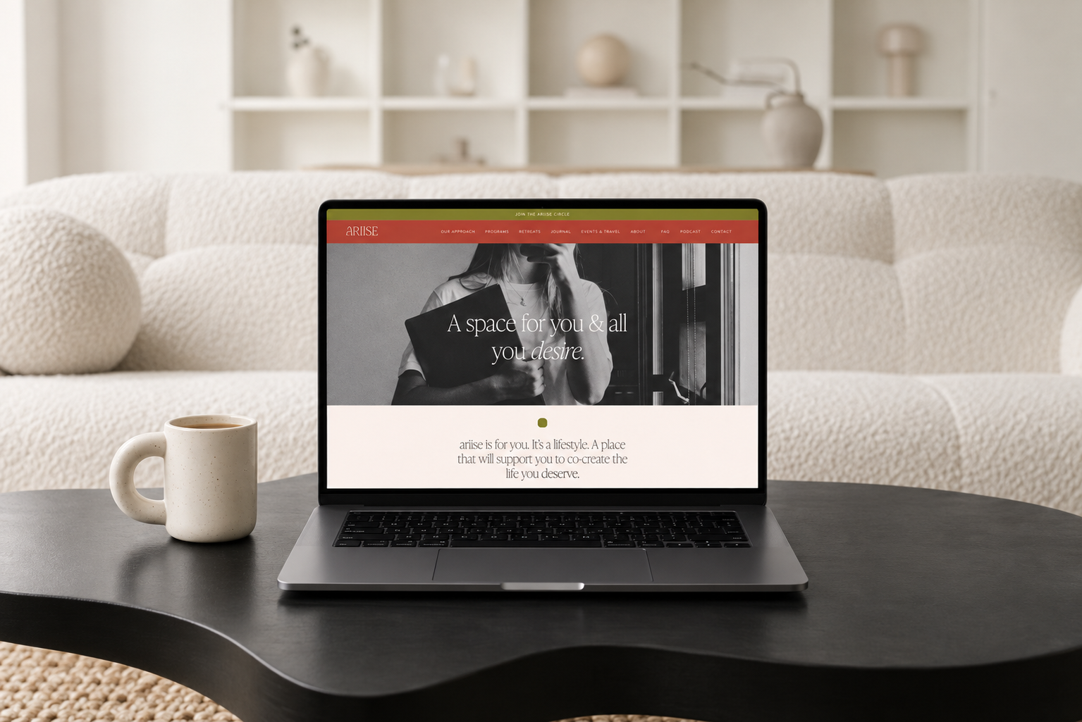 Laptop on coffee table displaying a website with the headline 'A space for you & all you desire'. Coffee mug to the left of the laptop. Cream-colored sofa and shelves with decorative items in the background.