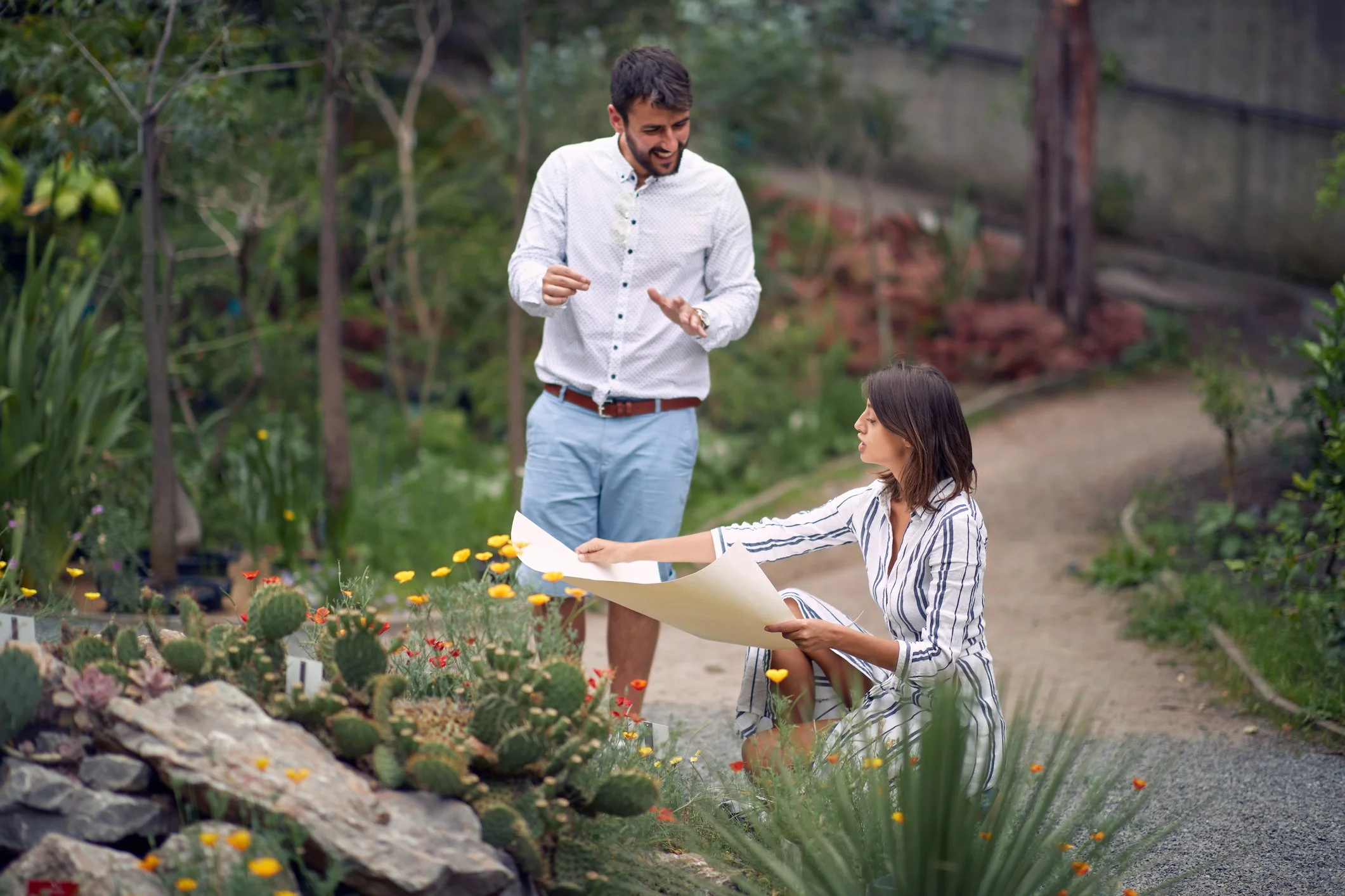 A man and woman in a garden discussing a large piece of paper or blueprint among desert plants, including cacti and succulents.