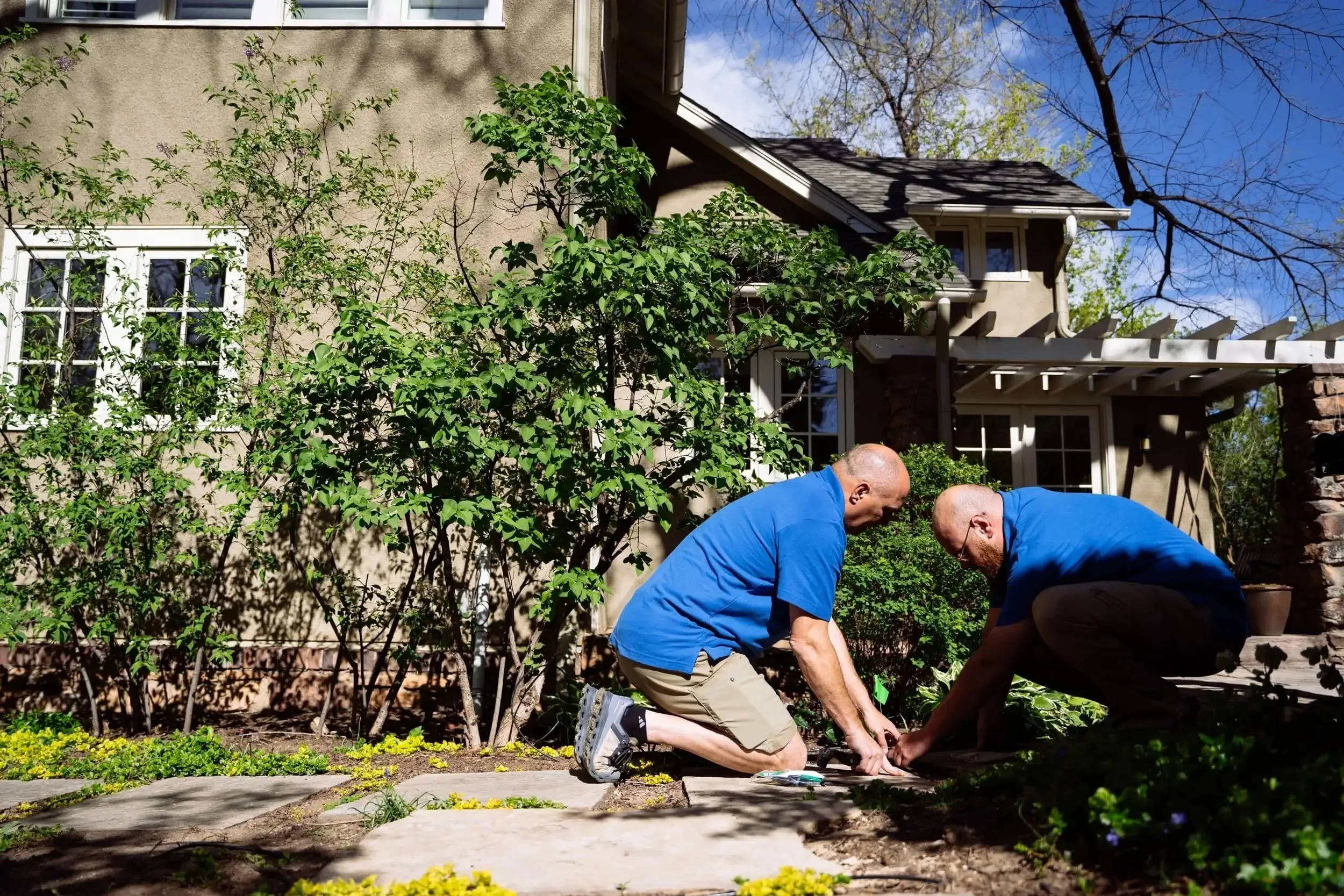 Two men in blue shirts kneeling on a garden path, working together in front of a house with beige walls and white-framed windows, planting or repairing plants on a sunny day.