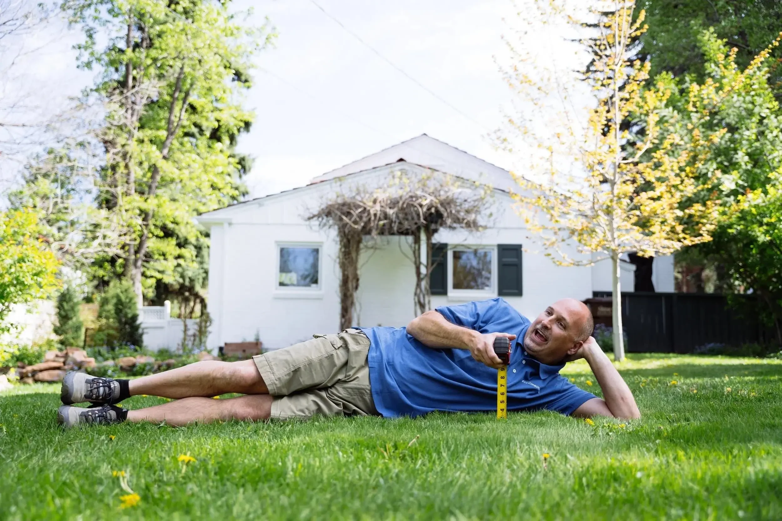 A man lying on the grass in a backyard, measuring the grass height with a ruler, with a white house and trees in the background.