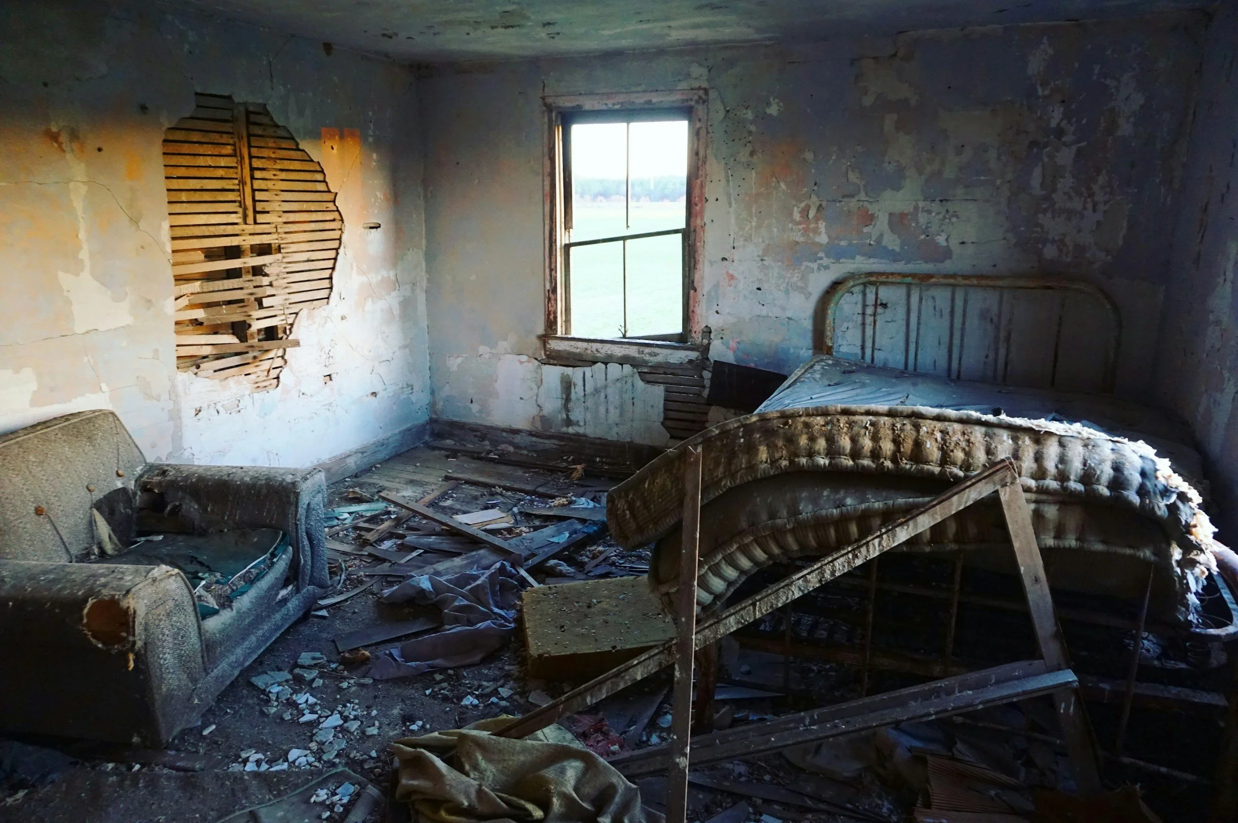 An abandoned, dilapidated bedroom with peeling paint, torn furniture, and debris scattered on the floor, illuminated by natural light from a window.