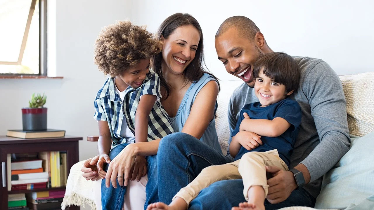Family of four smiling and sitting together on a sofa in a bright living room.