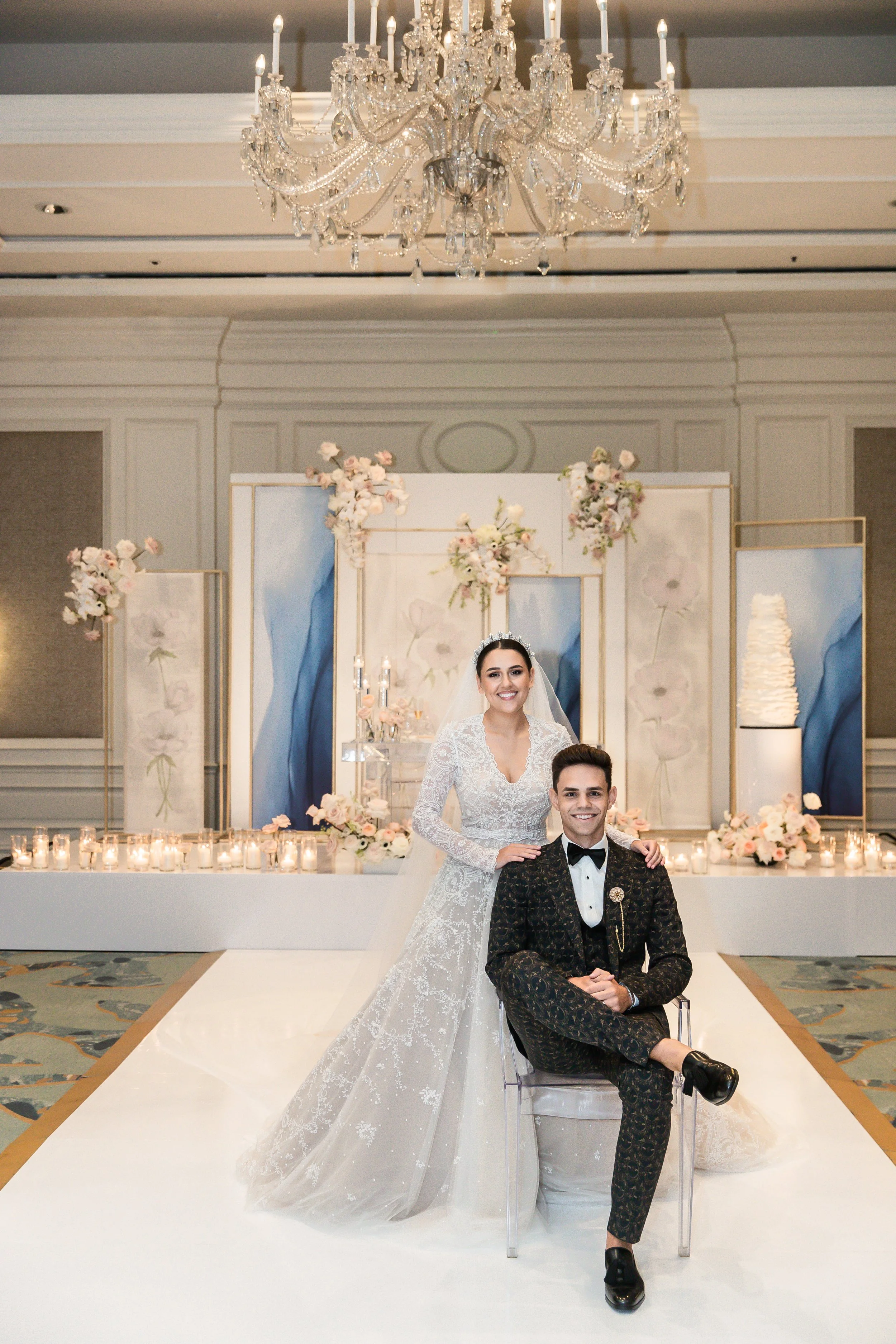 A bride and groom posing at their wedding in a decorated ballroom with a chandelier and floral arrangements.