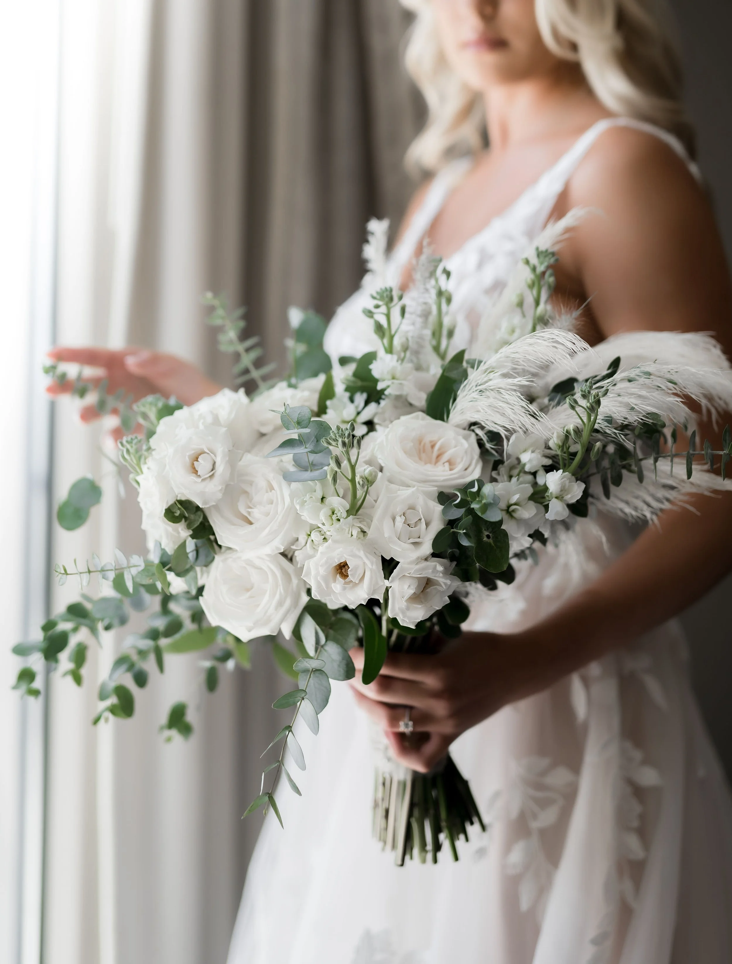 Woman holding a bouquet of white flowers and greenery in a wedding dress, standing near a window.