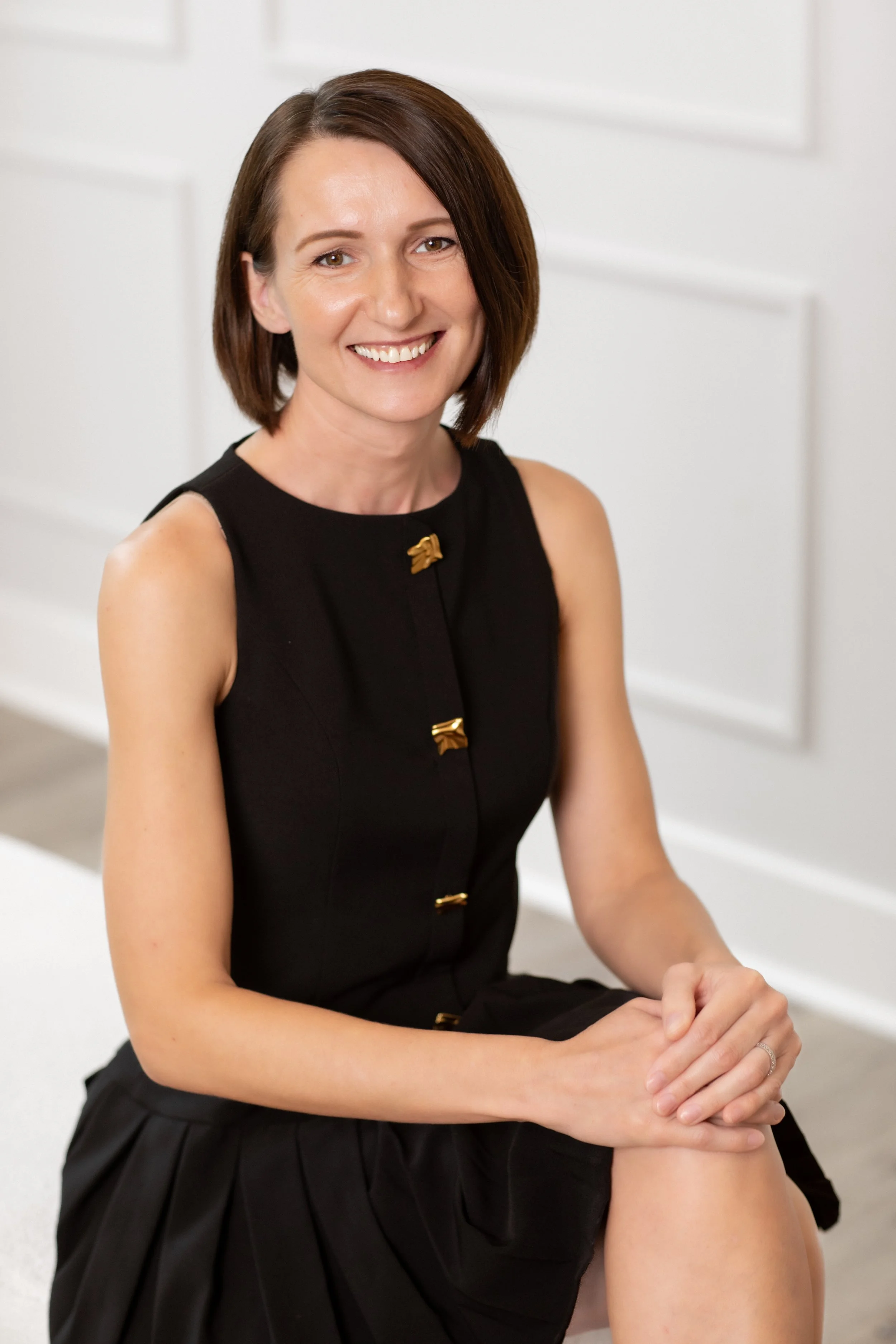 A woman with shoulder-length brown hair, wearing a sleeveless black dress with gold buttons, sitting on a white surface, smiling at the camera.