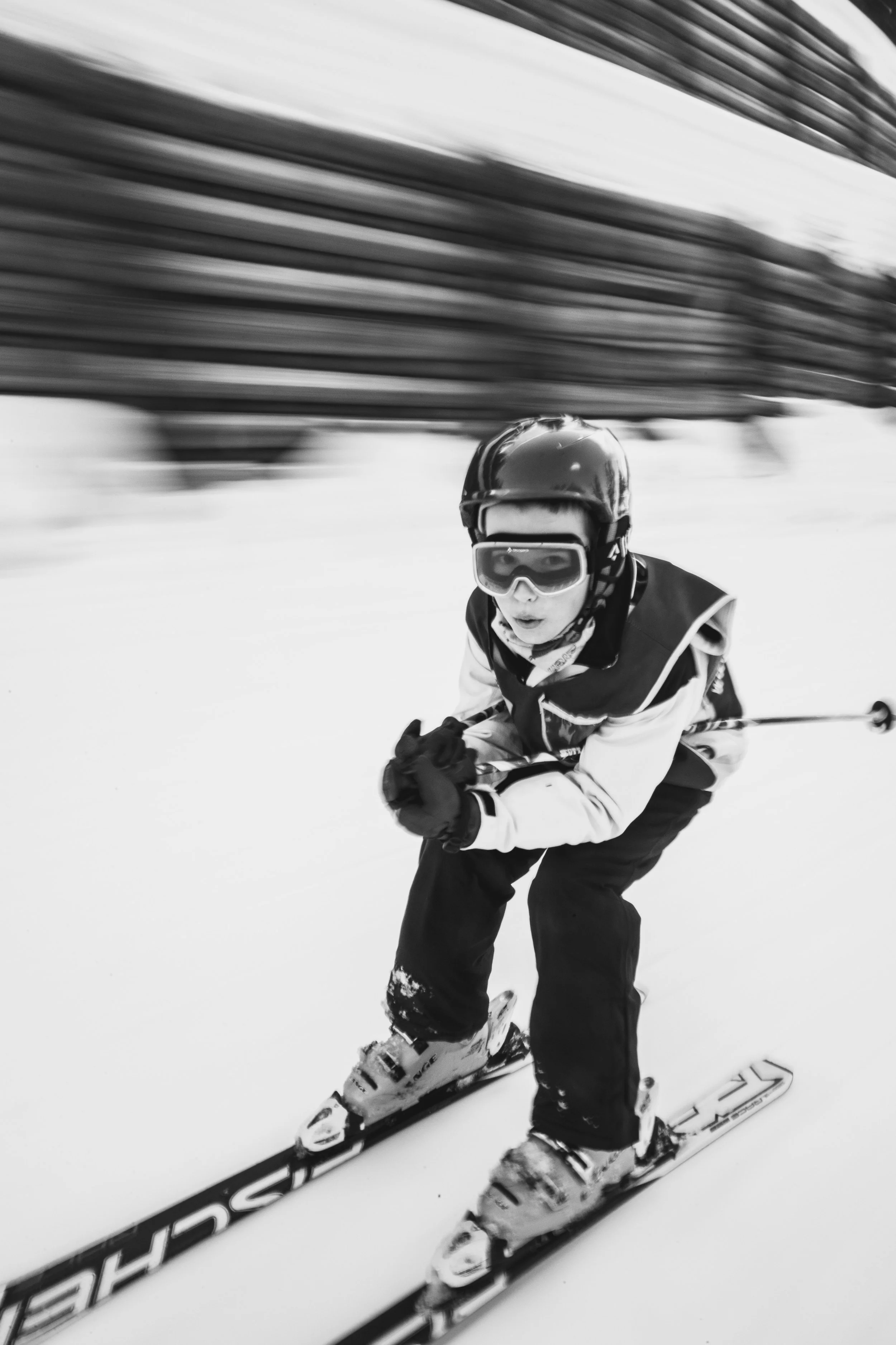 Black and white photo of a skier wearing a helmet, goggles, jacket, and gloves, skiing on snow with blurred buildings in the background.