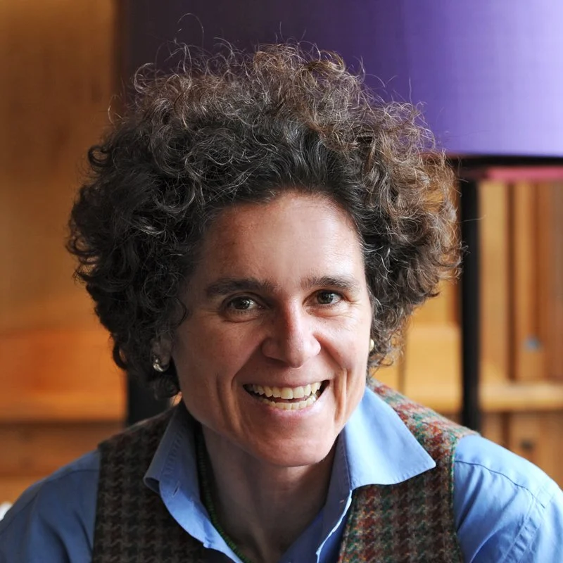 A woman with curly gray hair smiling at the camera, wearing a blue shirt and a plaid vest in a warmly-lit room.