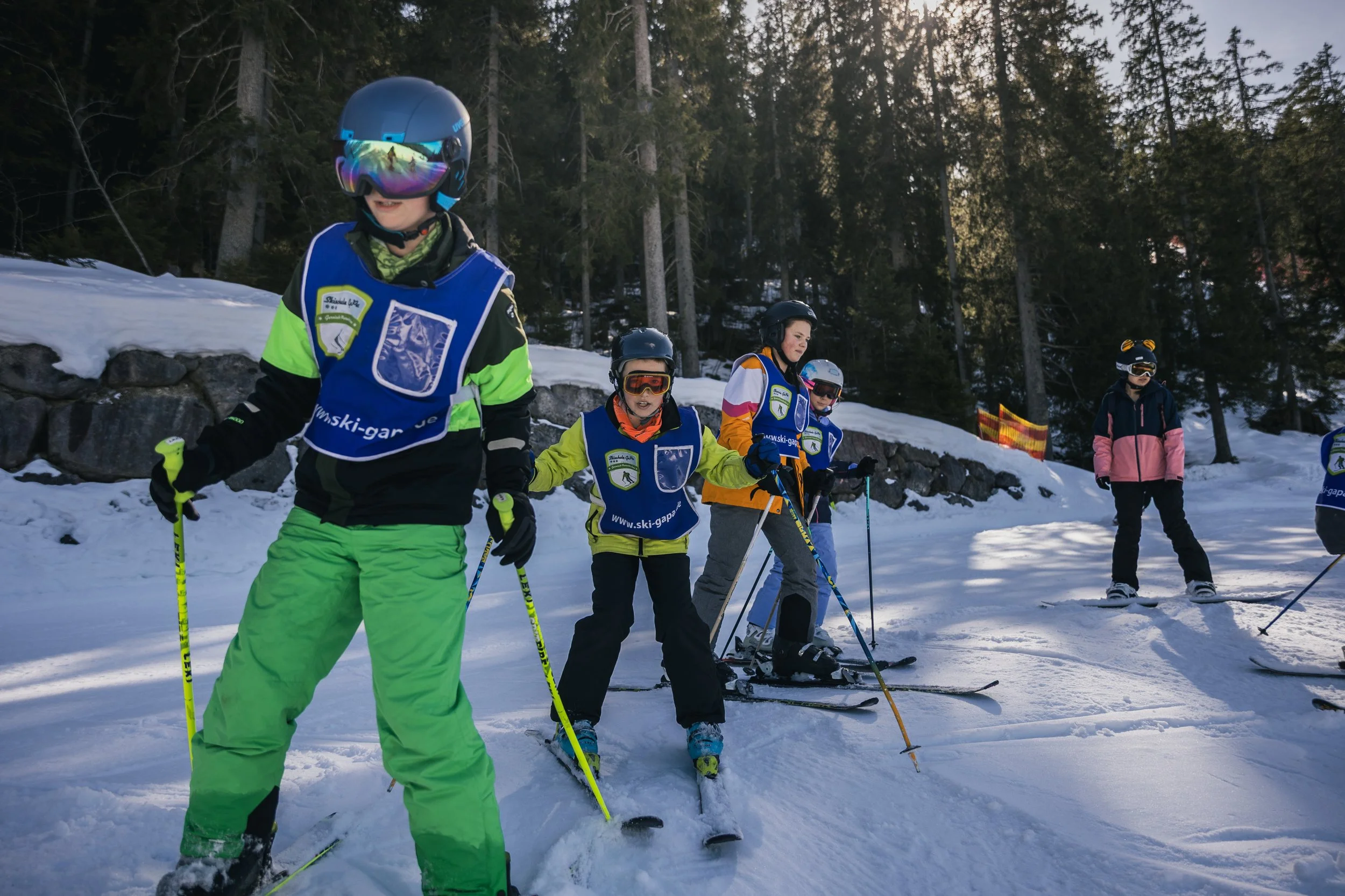 Children and an adult on skis learning on a snowy slope surrounded by tall trees, with children wearing blue vests with a logo, helmets, and winter gear.