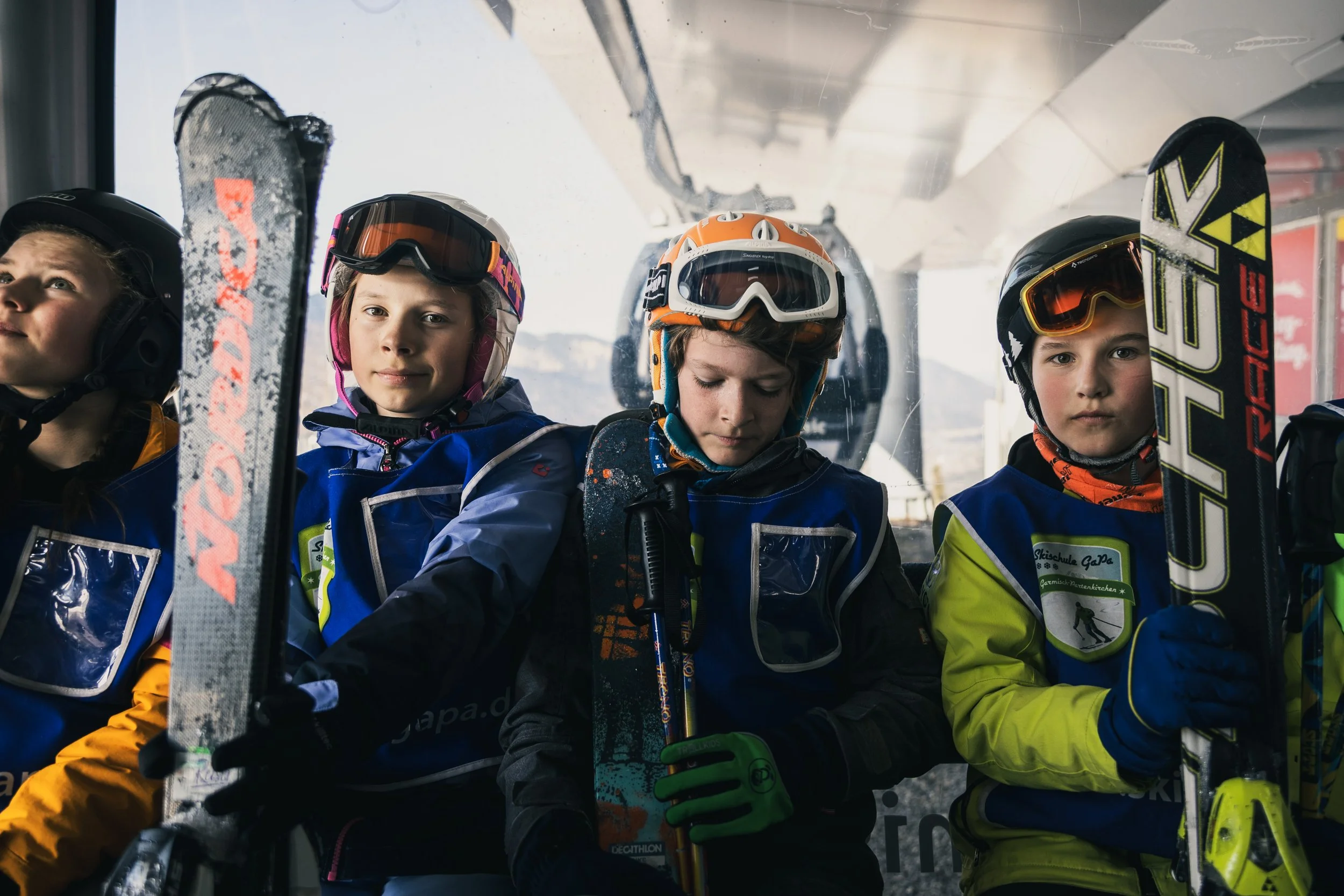 Four children in ski gear sitting in a cable car, holding ski equipment and wearing helmets and goggles.