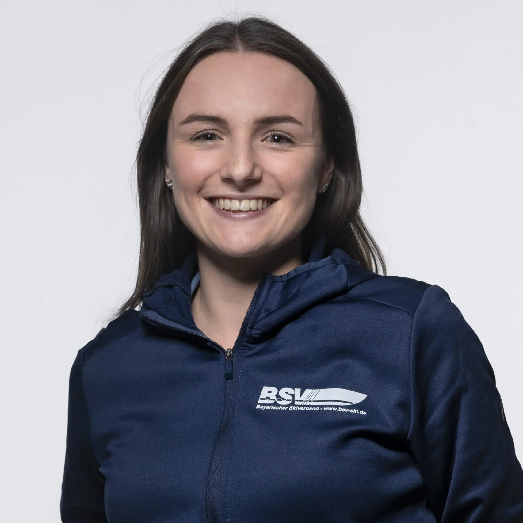 A smiling woman with long brown hair wearing a navy blue jacket with a logo on the chest that says 'BSV' and 'Bayerischer Ski Verband'.