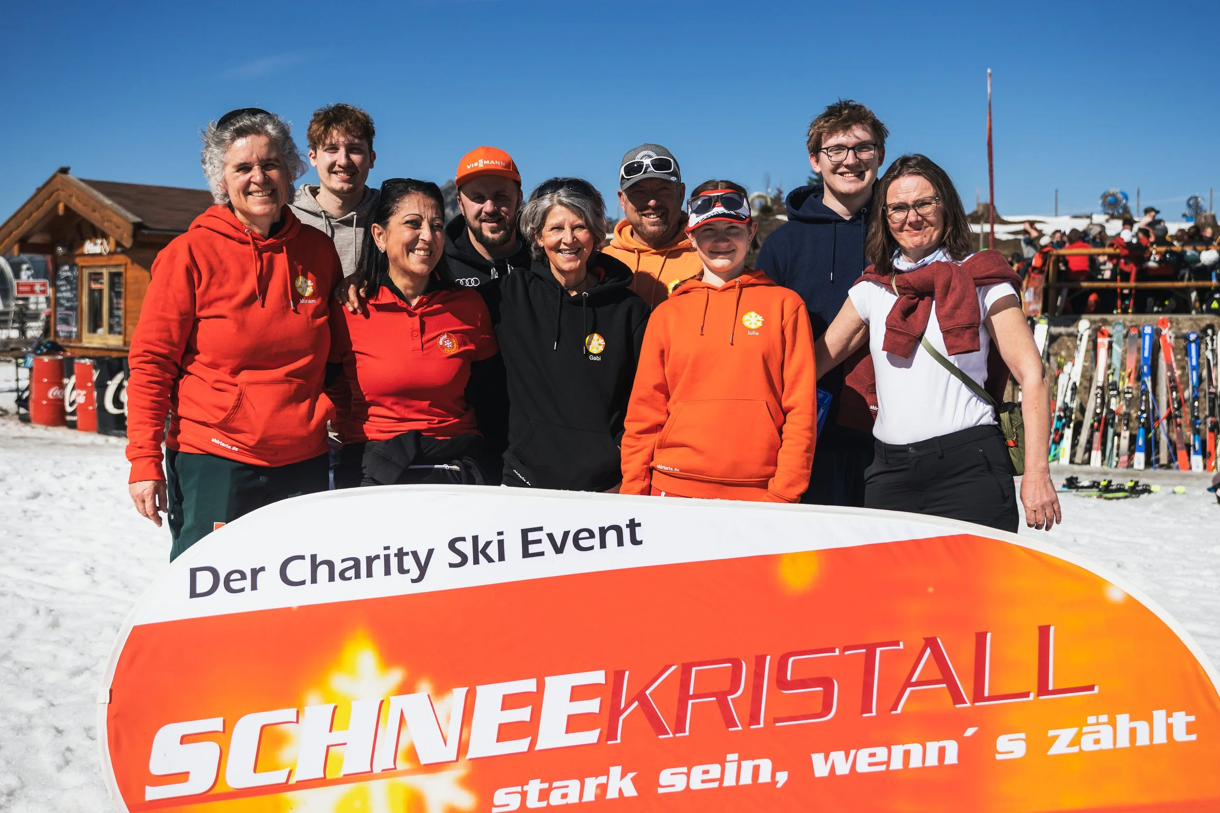 Group of people at a charity ski event, smiling and wearing colorful jackets, standing behind a banner with the text 'Der Charity Ski Event Schneekristall stark sein, wenn's zählt' on a snowy mountain.