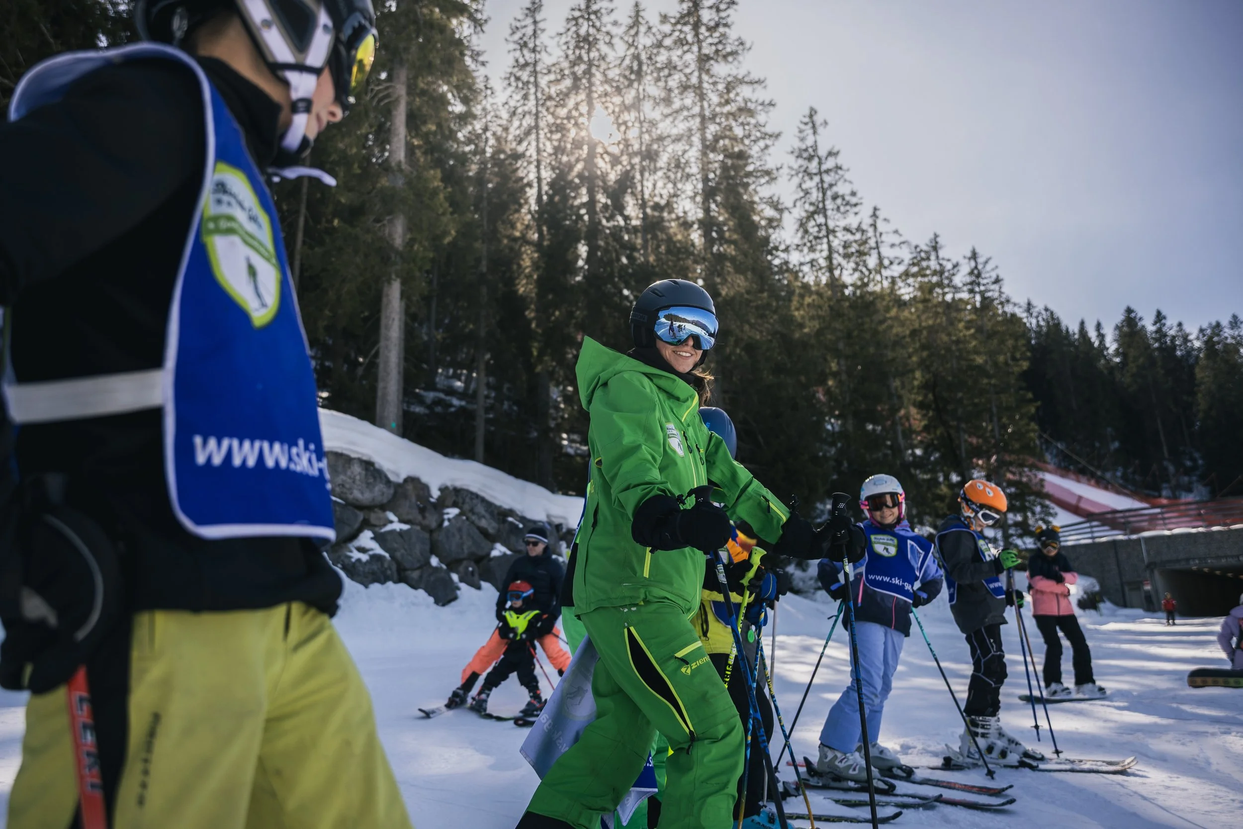 Group of skiers on a snowy mountain slope preparing to ski, with tall pine trees and a clear sky in the background.