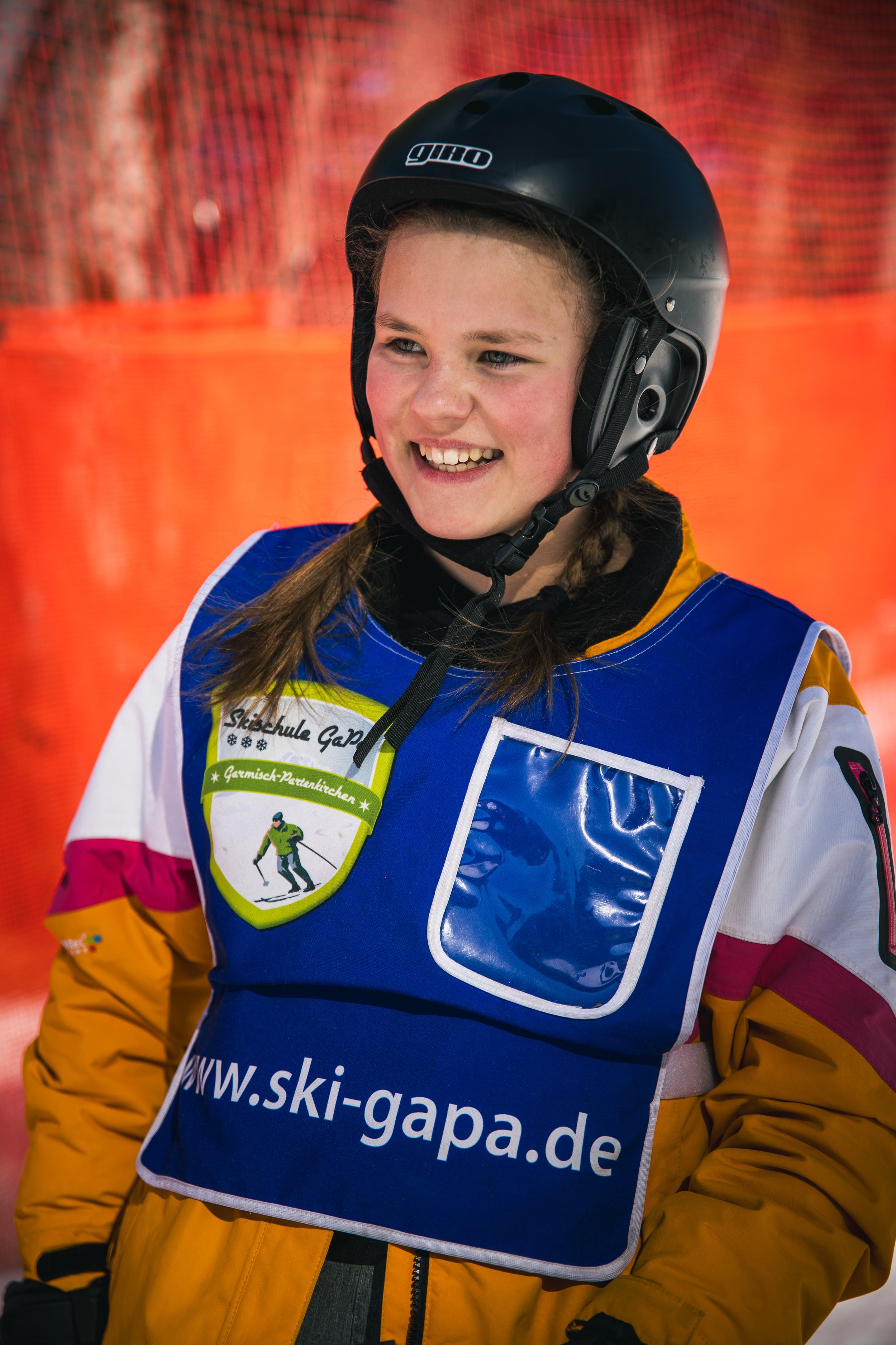 A young girl in snow ski gear, wearing a black helmet and a blue vest with a green and white patch and a website URL, smiling outdoors with orange fencing in the background.