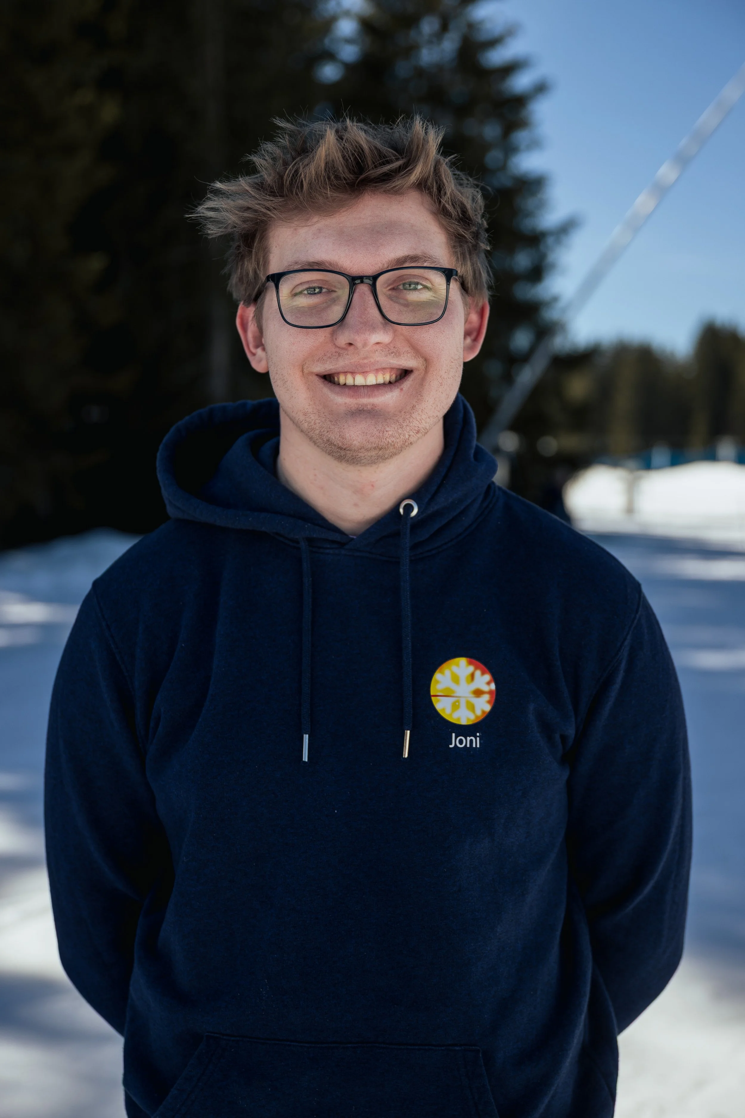 Young man smiling outdoors in winter, wearing glasses and a navy hoodie with a logo and the name 'Joni.'