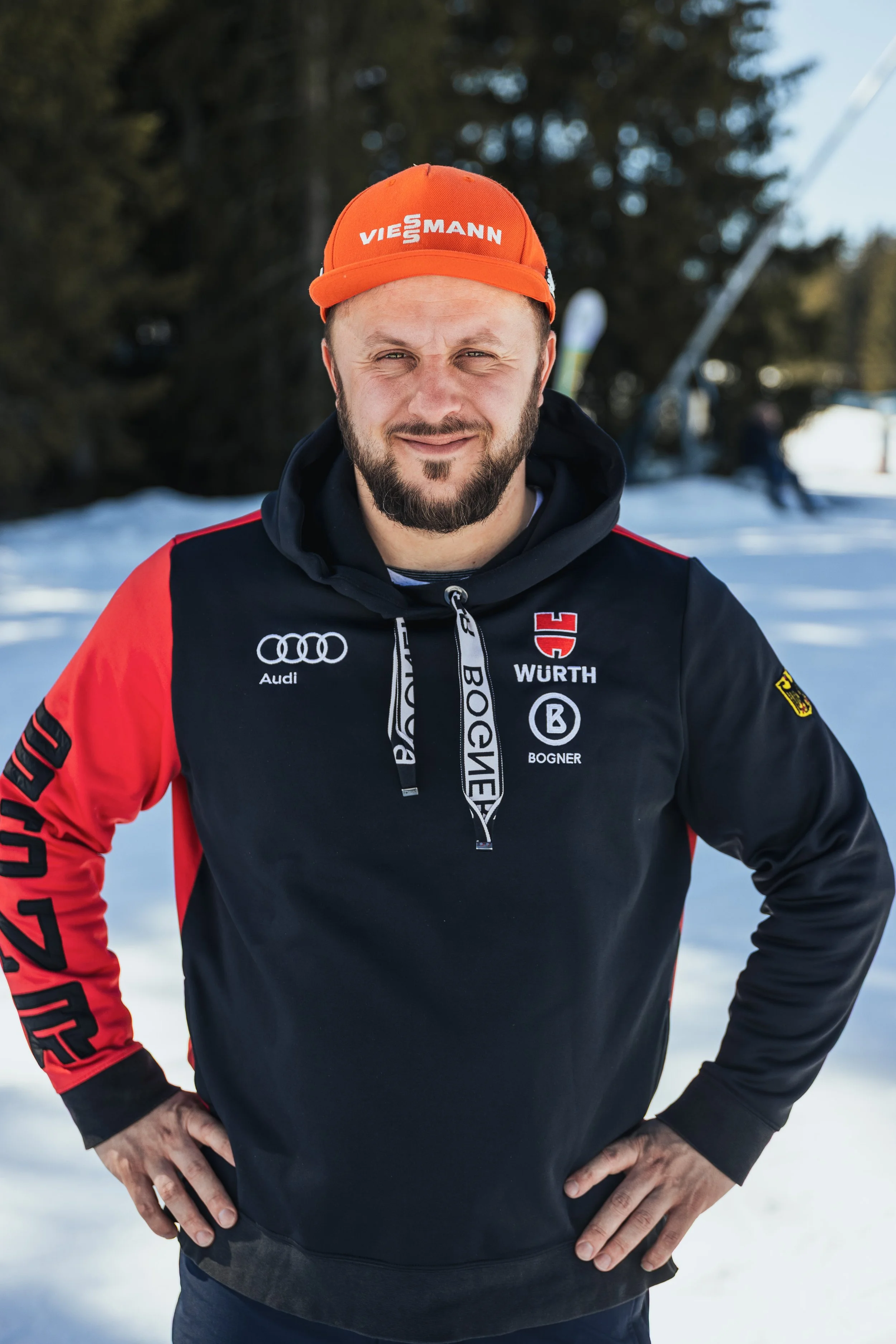 Man outdoors in winter snow, wearing a black and red sports jacket with sponsor logos, orange cap, and black hoodie.