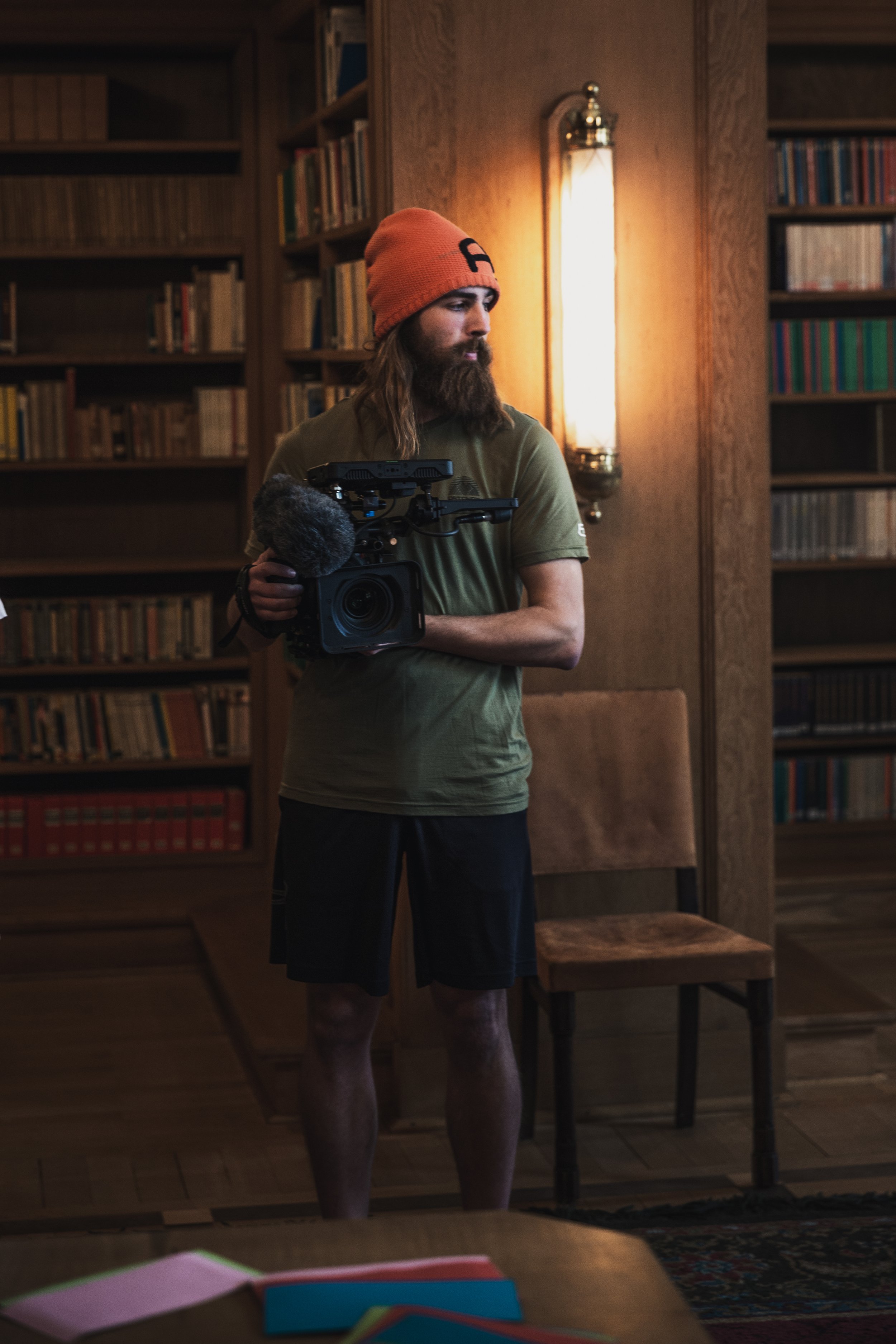 A man with long hair and a beard wearing a red beanie, olive green t-shirt, and black shorts holding a professional video camera in a wood-paneled room with bookshelves and a chair.