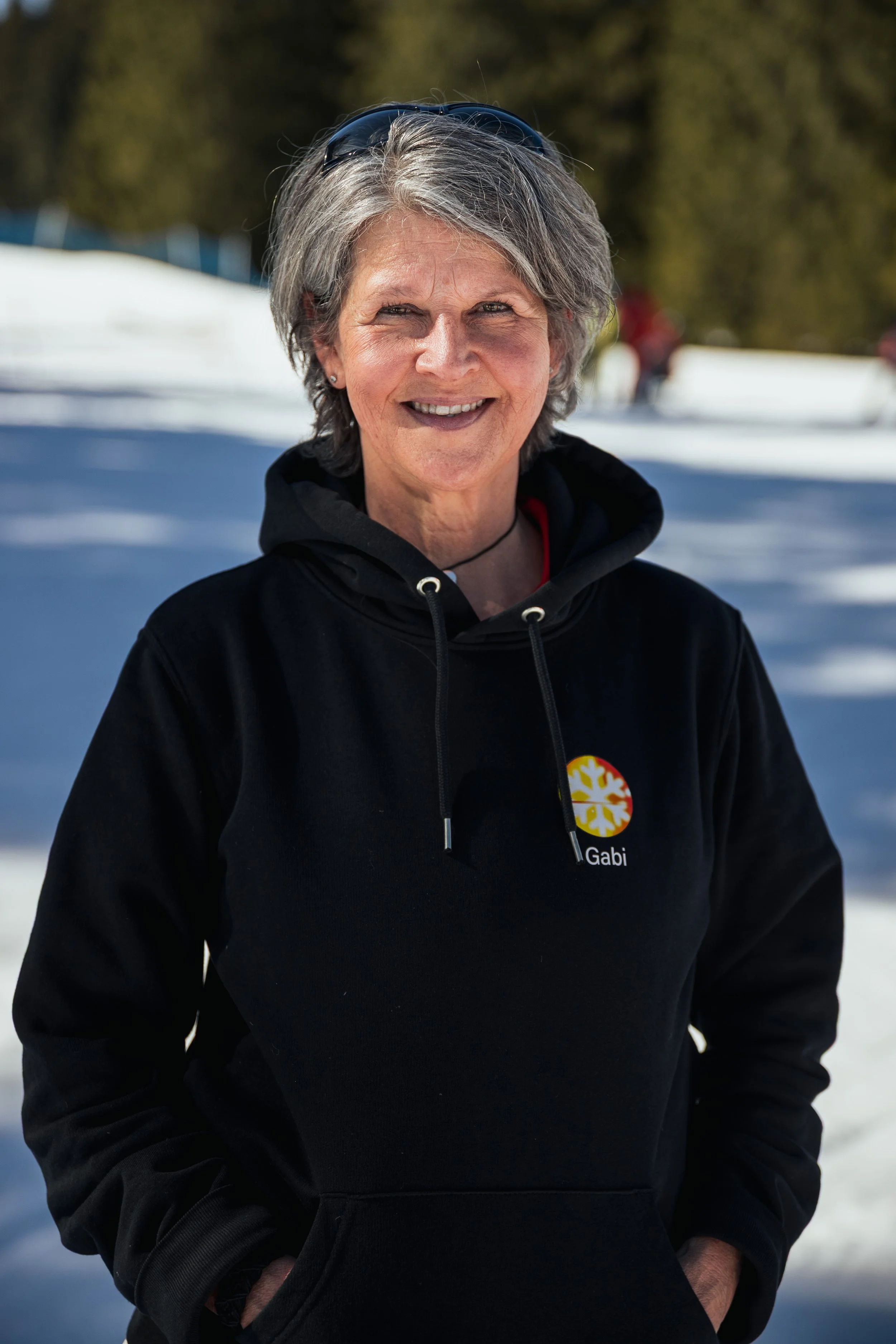 A smiling woman with short gray hair wearing sunglasses on her head and a black hoodie with a colorful logo and the name 'Gabi' printed on it, standing outdoors in a snowy landscape with trees in the background.