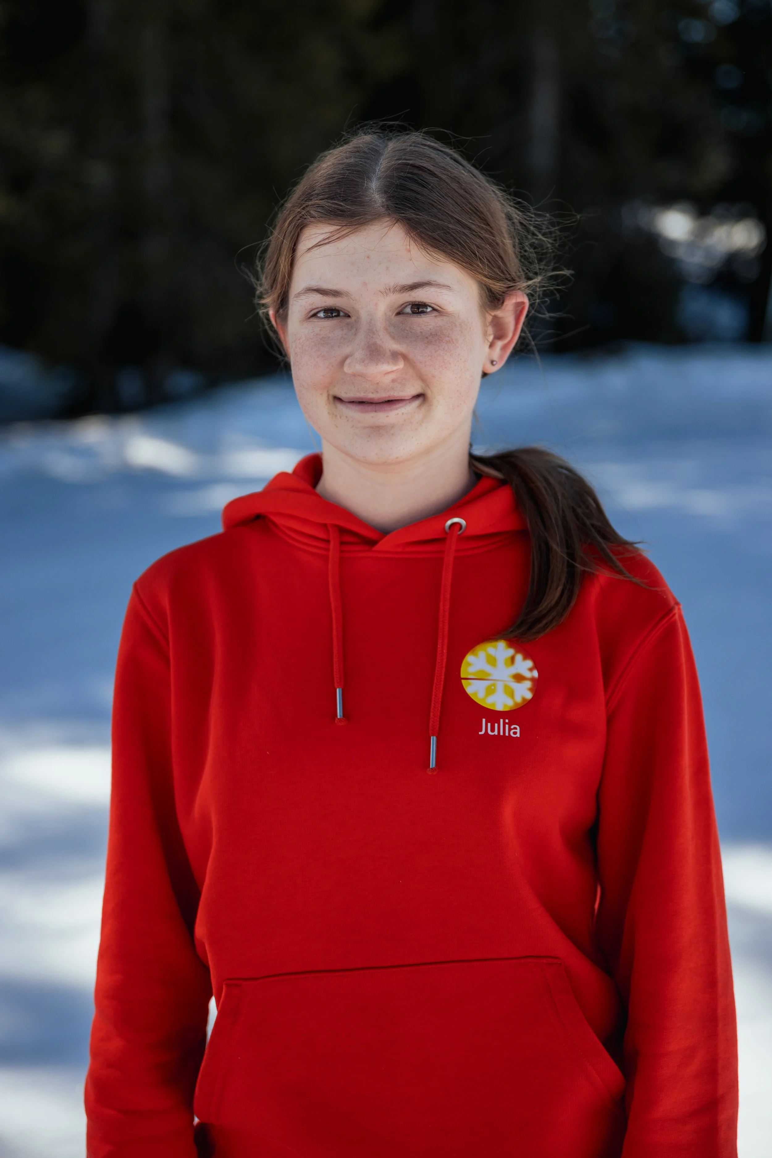 A young girl with freckles, wearing a red hoodie with a yellow and white snowflake logo and her name, Julia, on it, standing outdoors with snow and trees in the background.